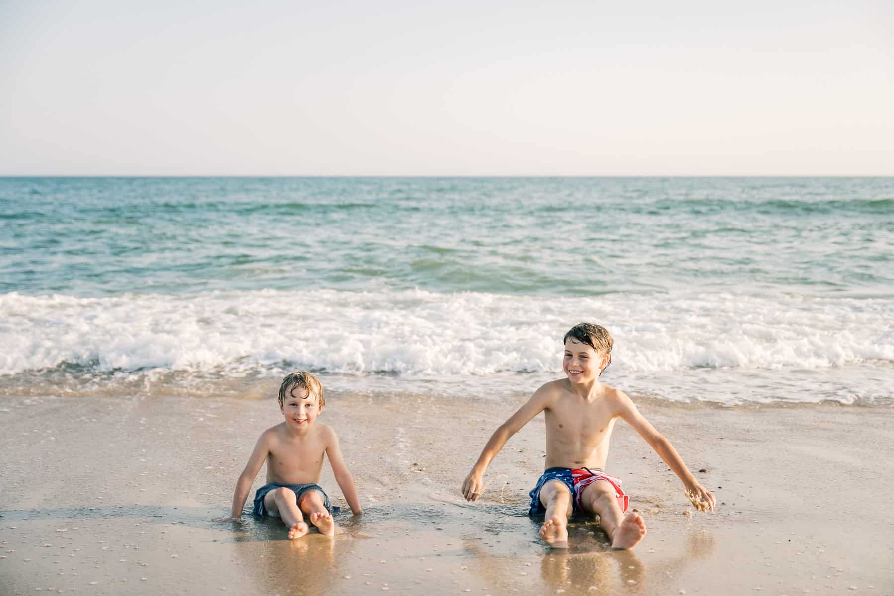 Two young boys in swim trunks sit on wet sand at the ocean’s edge, gentle waves behind them. Captured by Nantucket photographer Kaity Farrell, this scene is perfect for a memorable family photo session.