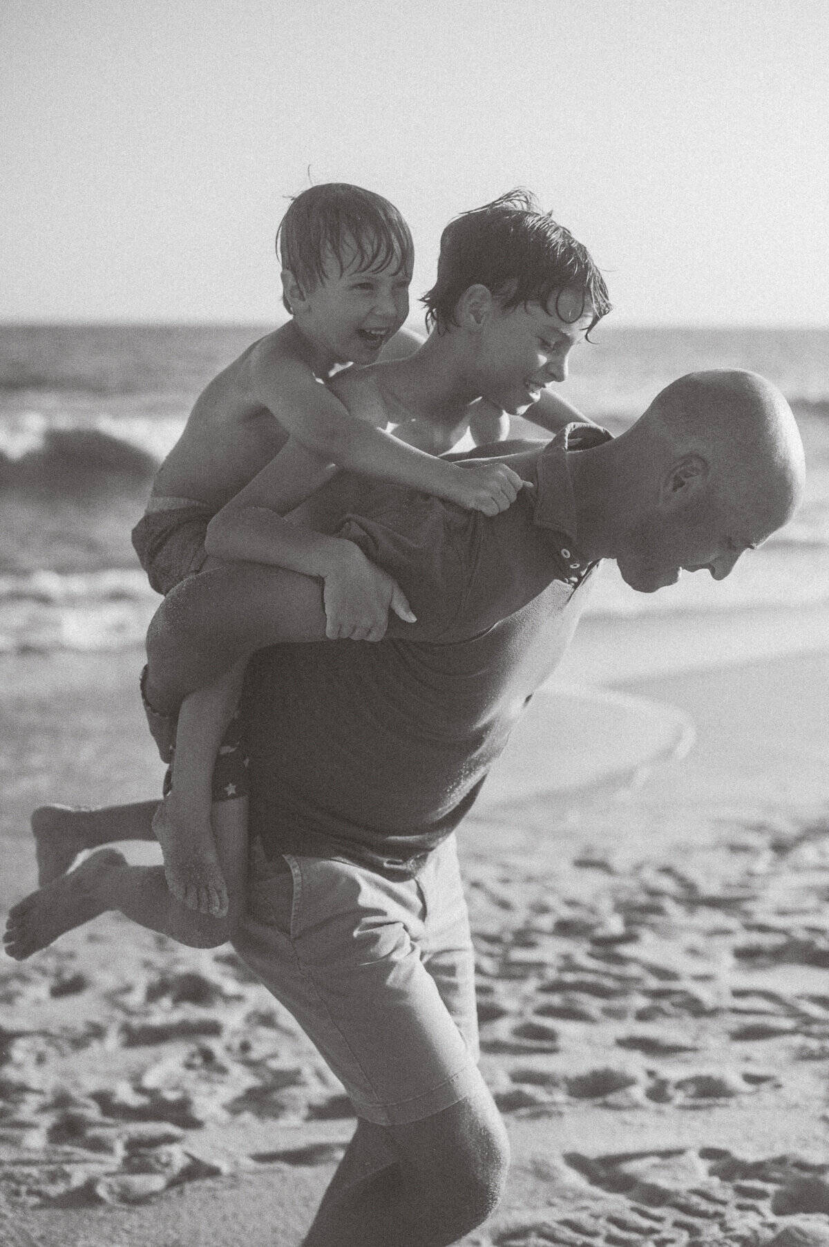 A man carries two young boys on his back along a sandy beach near the ocean, captured by Nantucket Family Portrait Photographer Kaity Farrell.