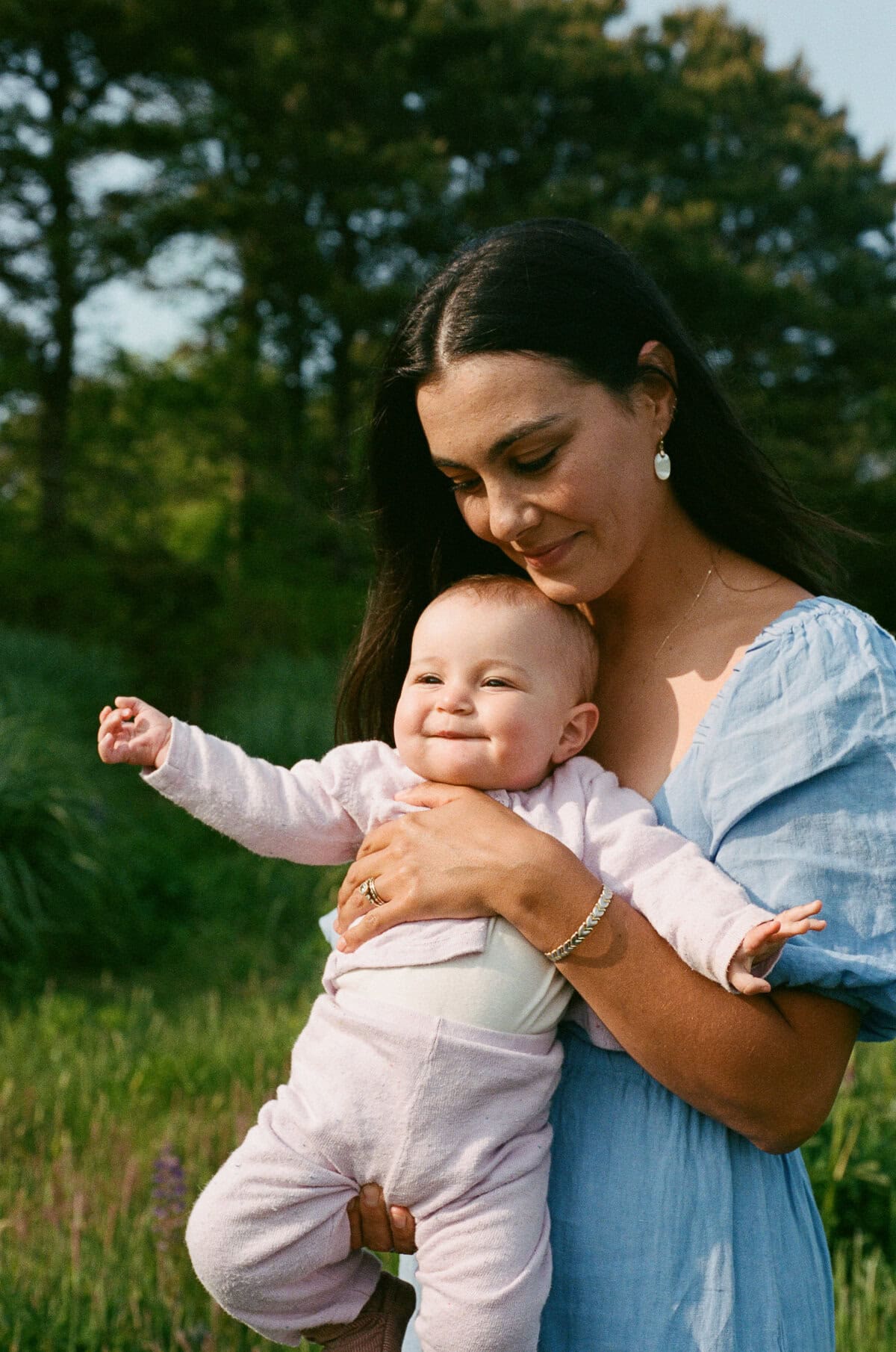 A woman in a blue dress holds a smiling baby in a light purple outfit outdoors with trees and grass, captured during a family photo session by Nantucket photographer Kaity Farrell.
