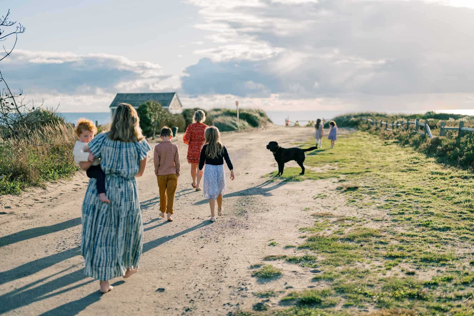 A group of people, including children and a dog, walk barefoot on a dirt path toward Madaket beach in Nantucket on a sunny day during a family portrait session with Nantucket photographer Kaity Farrell.