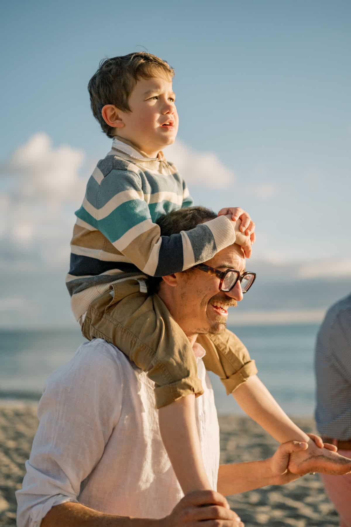 A child sits on an adult’s shoulders at the beach in Nantucket, both looking into the distance—captured during a Nantucket family portrait session by Nantucket photographer Kaity Farrell. The adult wears glasses and a white shirt; the child wears a striped sweater and rolled-up pants.
