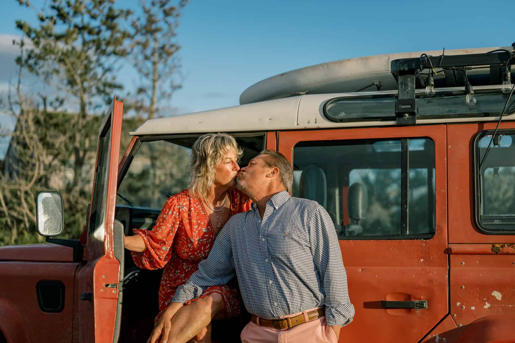 A couple kisses next to an open door of a red off-road vehicle parked outdoors on a sunny day, captured during a couples photo session by Nantucket photographer Kaity Farrell.