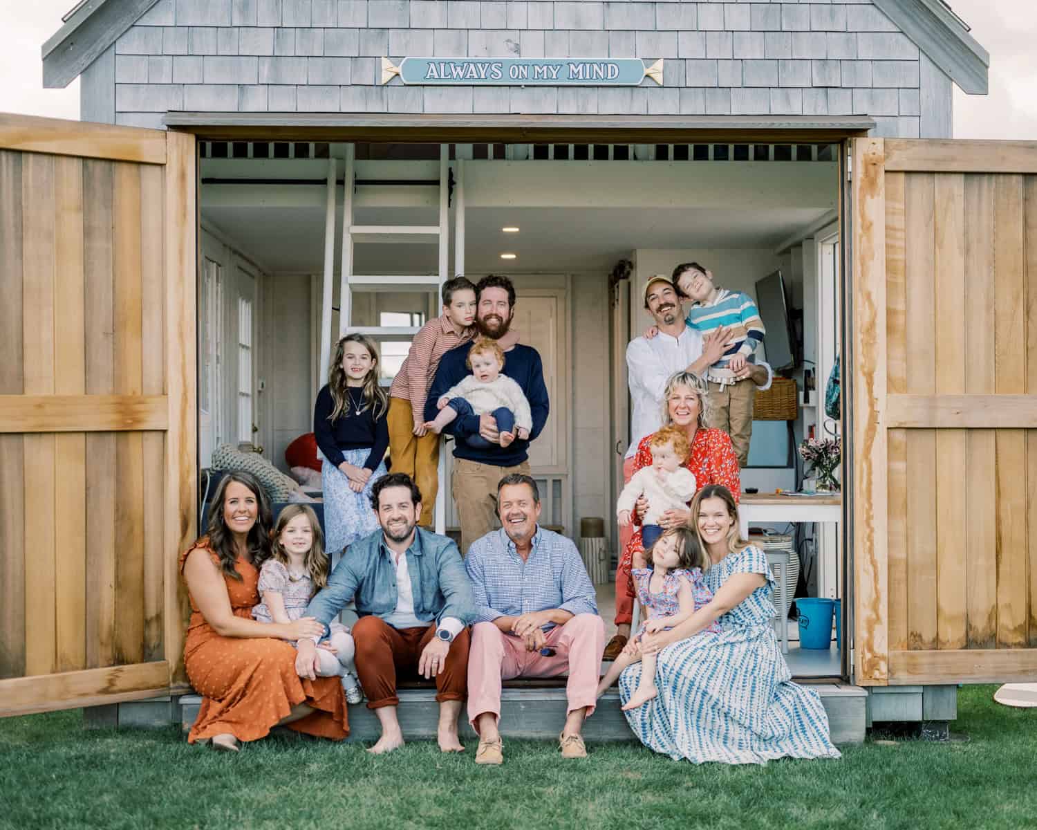 A multigenerational family of twelve people, including adults and children, pose for a photo in front of an open wooden shed with a sign reading "Always on My Mind," captured by Nantucket portrait photographer Kaity Farrell.