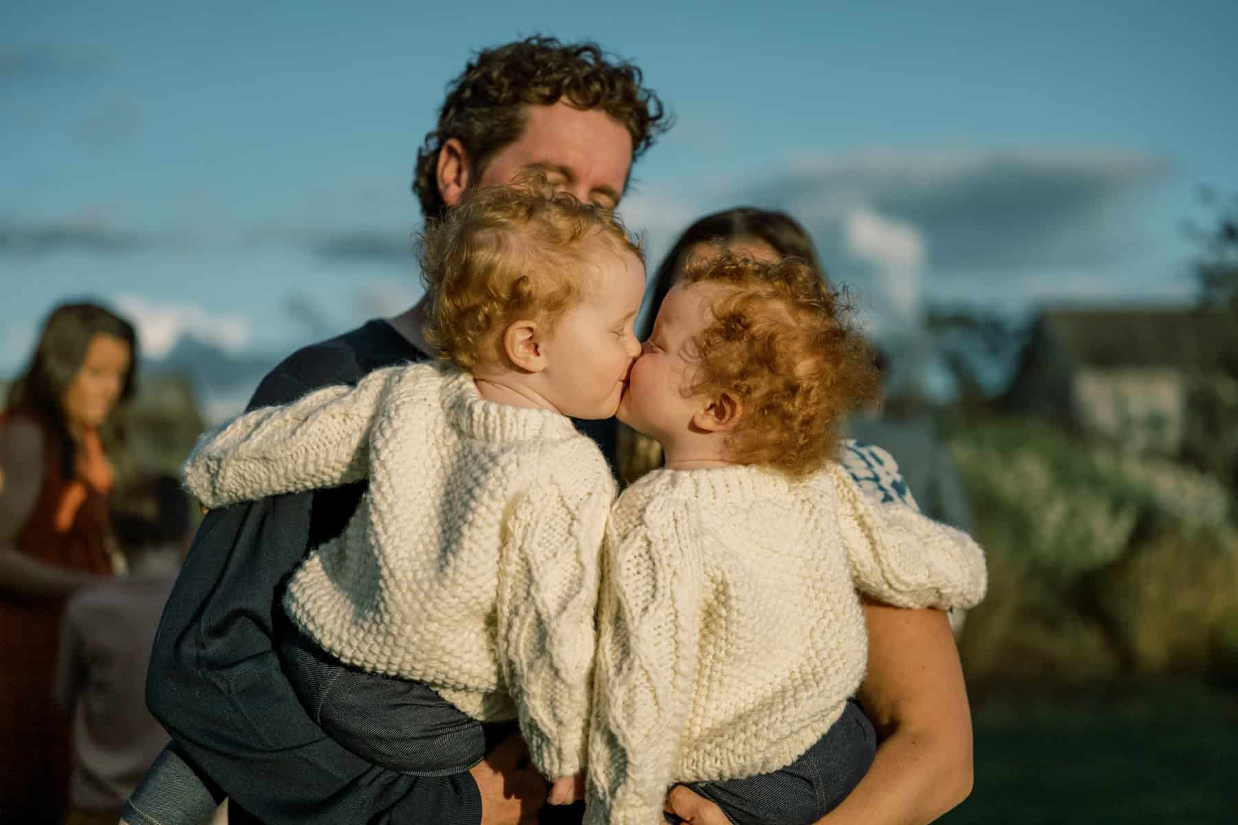 Two toddlers with curly hair and matching cream sweaters kiss each other while being held by adults outdoors on a sunny day during a family photo session with Nantucket photographer Kaity Farrell.