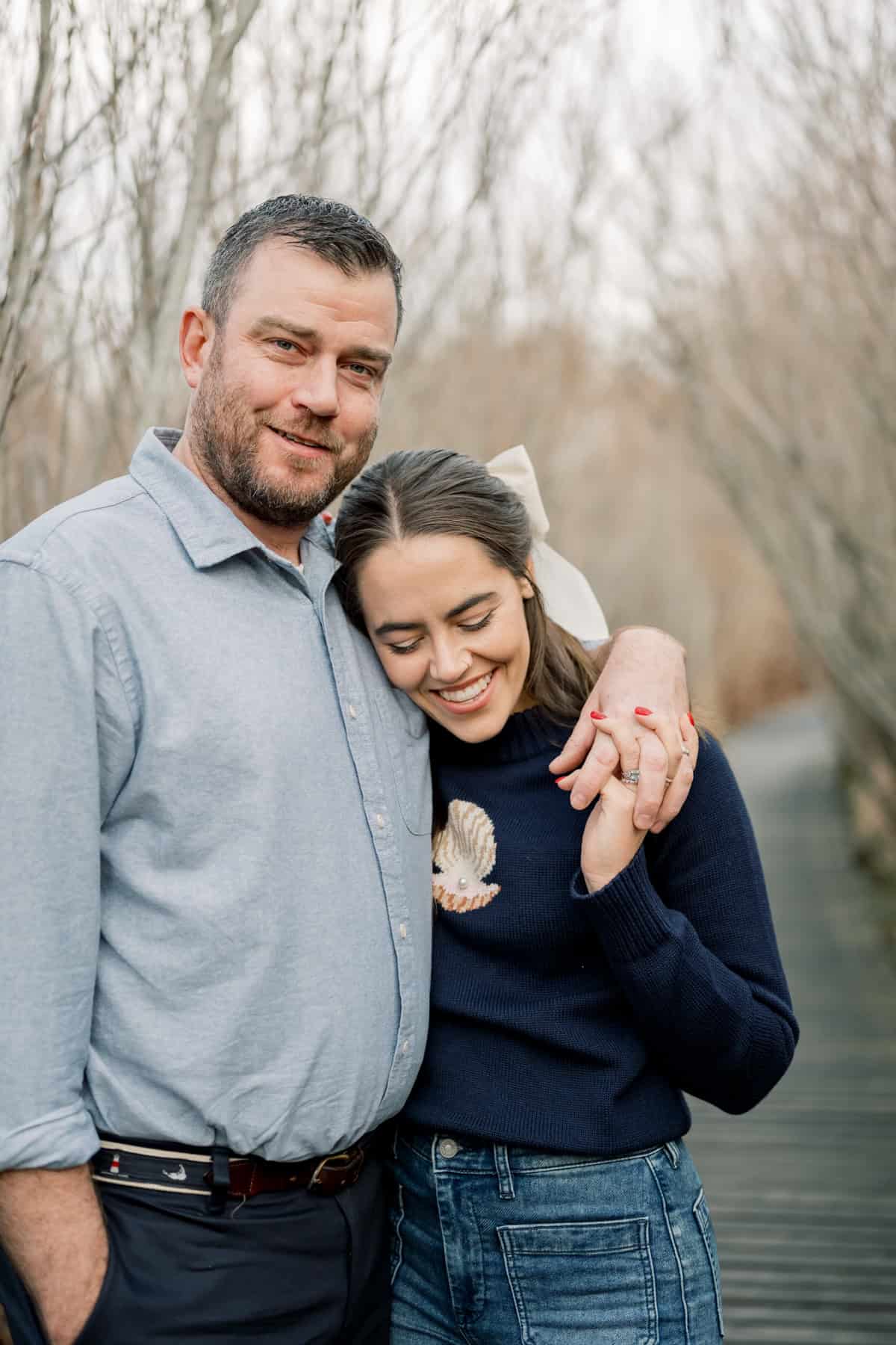 A man with his arm around a smiling woman who is leaning on his shoulder, standing on an outdoor path lined with bare trees—captured during a couples portrait session by Nantucket photographer Kaity Farrell.