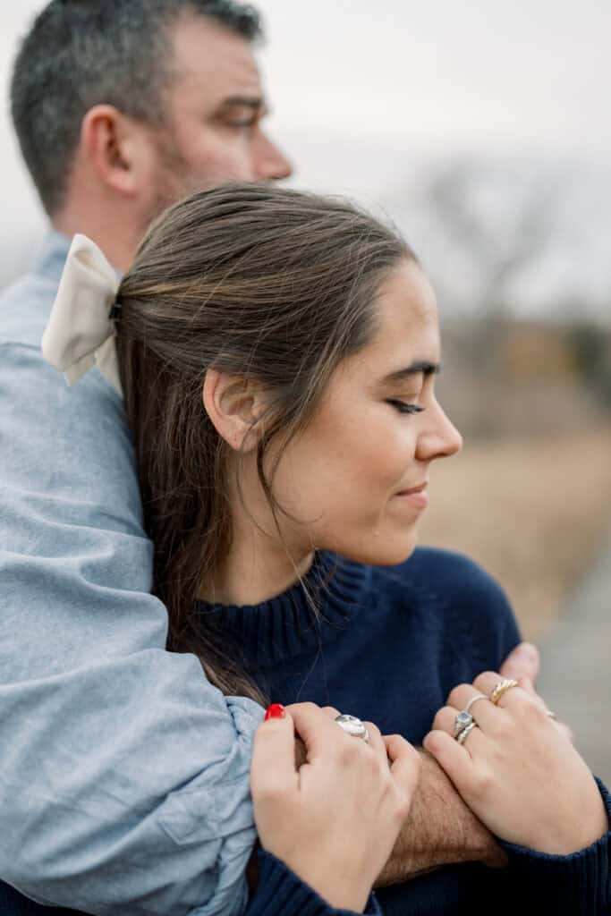 A woman with closed eyes holds hands with a man embracing her from behind. Both wear blue; the woman has a white hair bow and rings. Captured during a couples portrait session by Nantucket photographer Kaity Farrell.