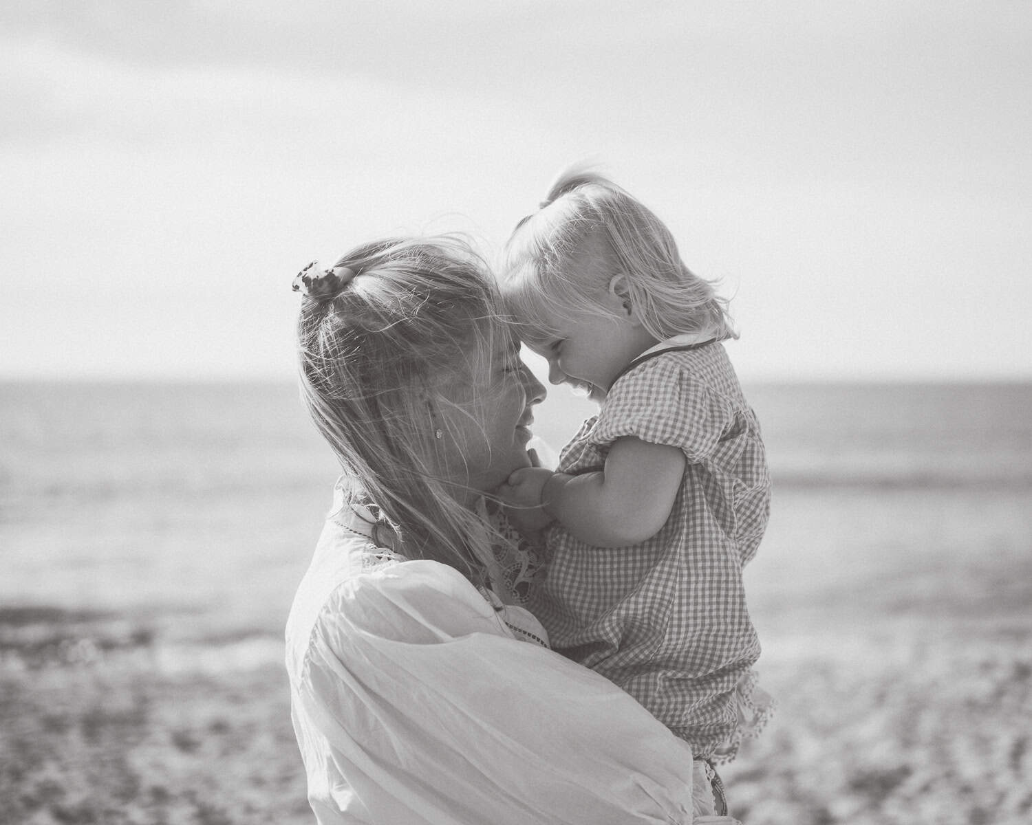 A woman holding a young child at the beach; they touch their foreheads together and smile. Captured by Nantucket photographer Kaity Farrell, this black and white image beautifully reflects the warmth of a Nantucket family moment.