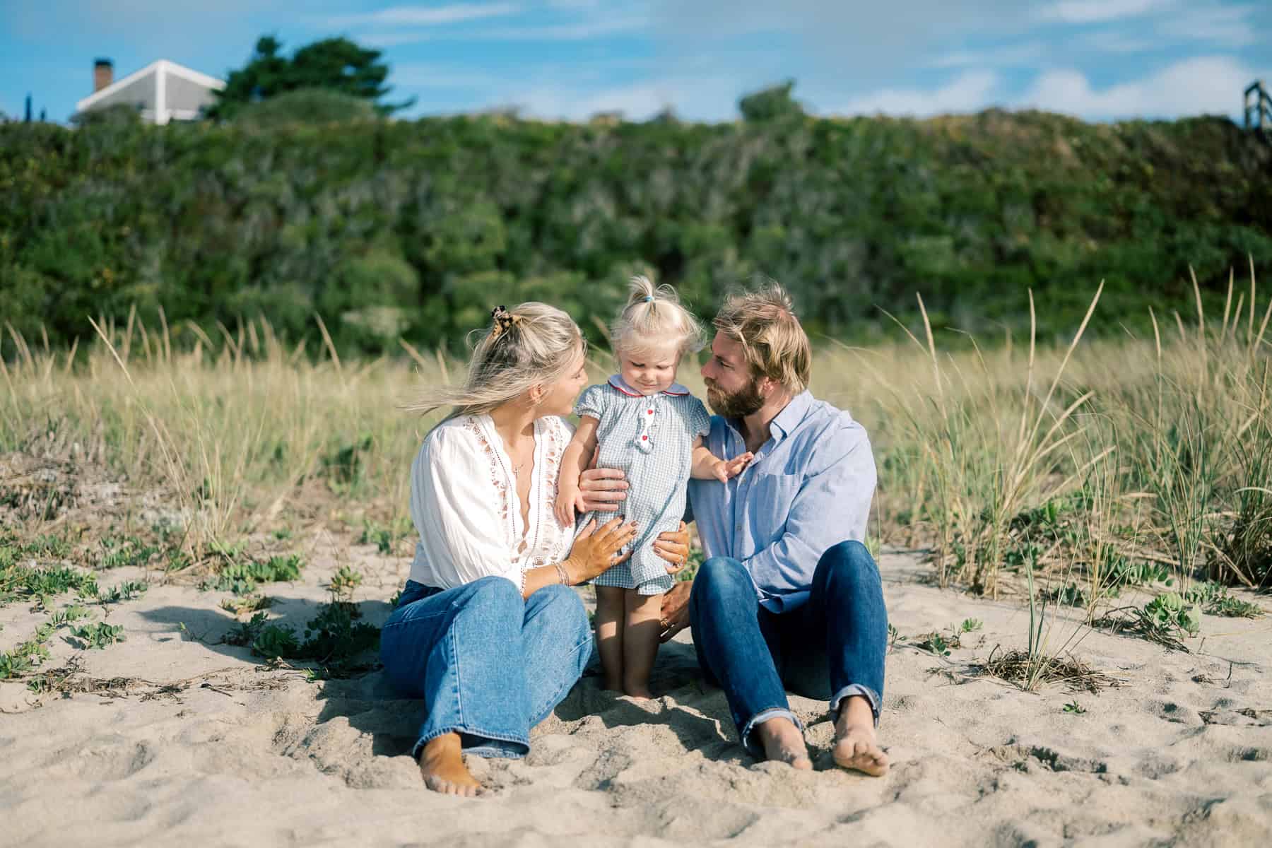 A man, woman, and young child sit together on sand at the beach, surrounded by tall grass with a house and greenery in the background—captured beautifully by Nantucket Family Portrait Photographer Kaity Farrell.