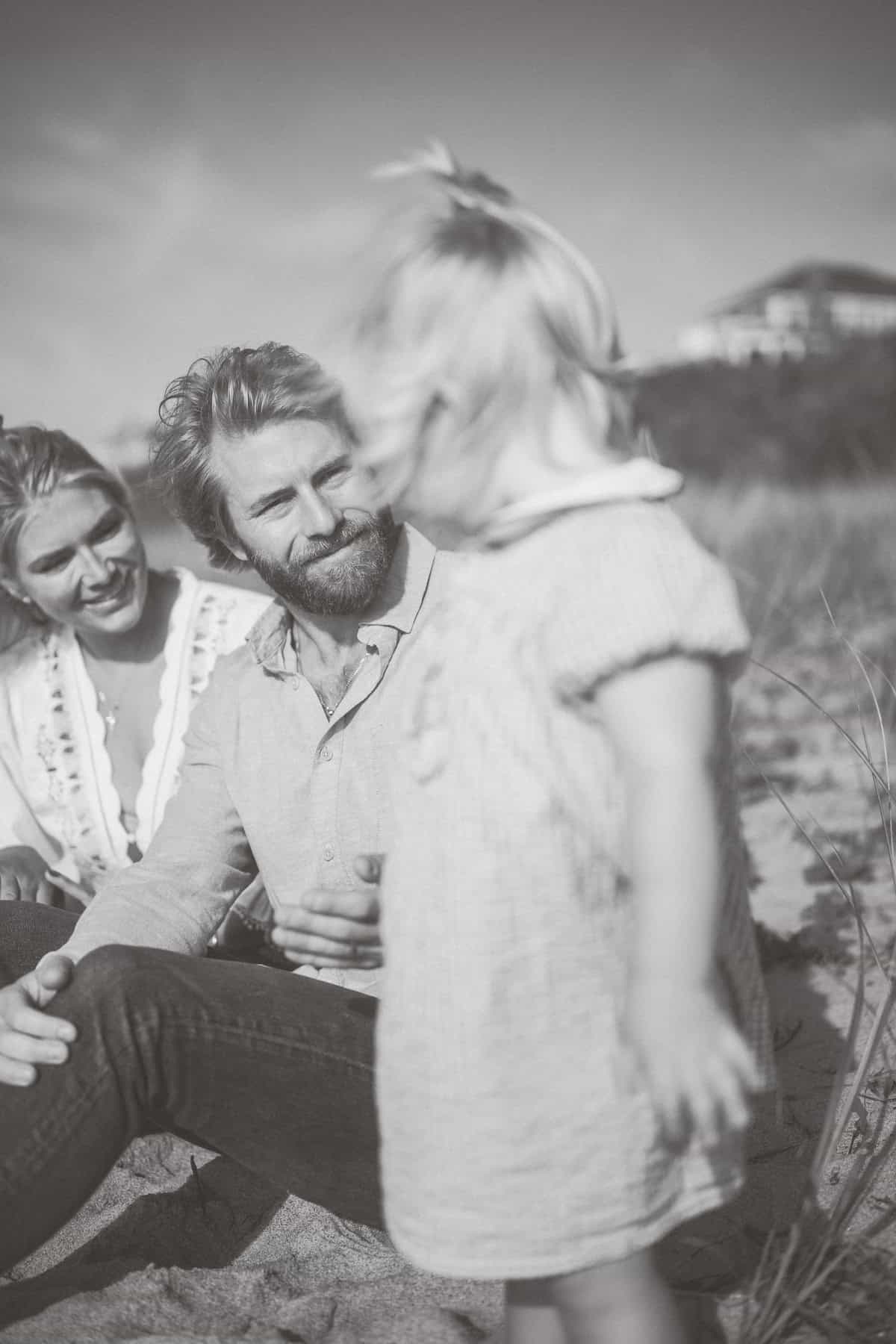 A man and woman sit on sand outdoors, gazing at a young child standing in the foreground. This black and white Nantucket family photo session was beautifully captured by Nantucket photographer Kaity Farrell.