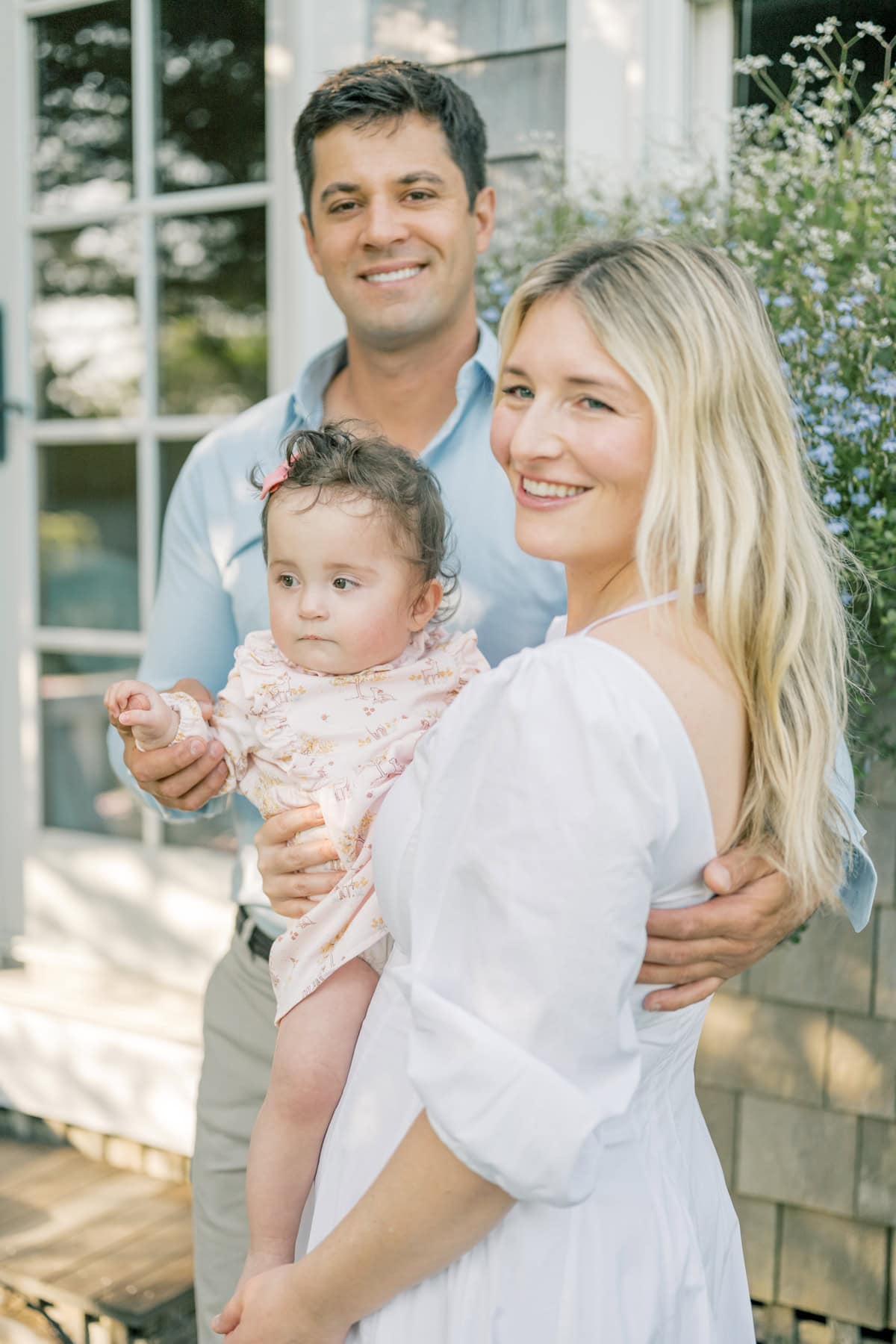 A man and woman stand outdoors smiling, holding a baby in front of a house with flowers visible in the background, captured by Nantucket family photographer Kaity Farrell.