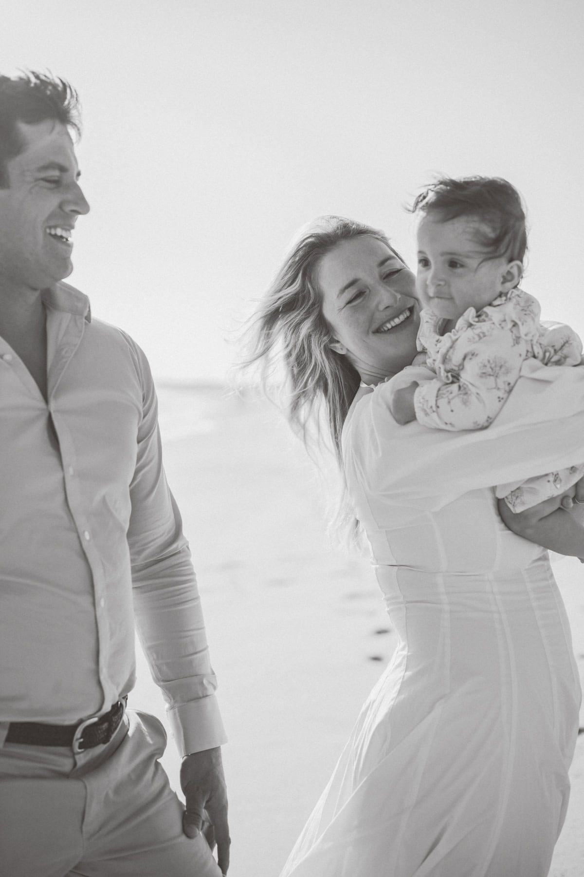 A man, woman, and baby stand together on a beach, smiling and looking happy. Captured in black and white, this Nantucket family enjoys a heartfelt moment during their family photo session with Nantucket photographer Kaity Farrell.