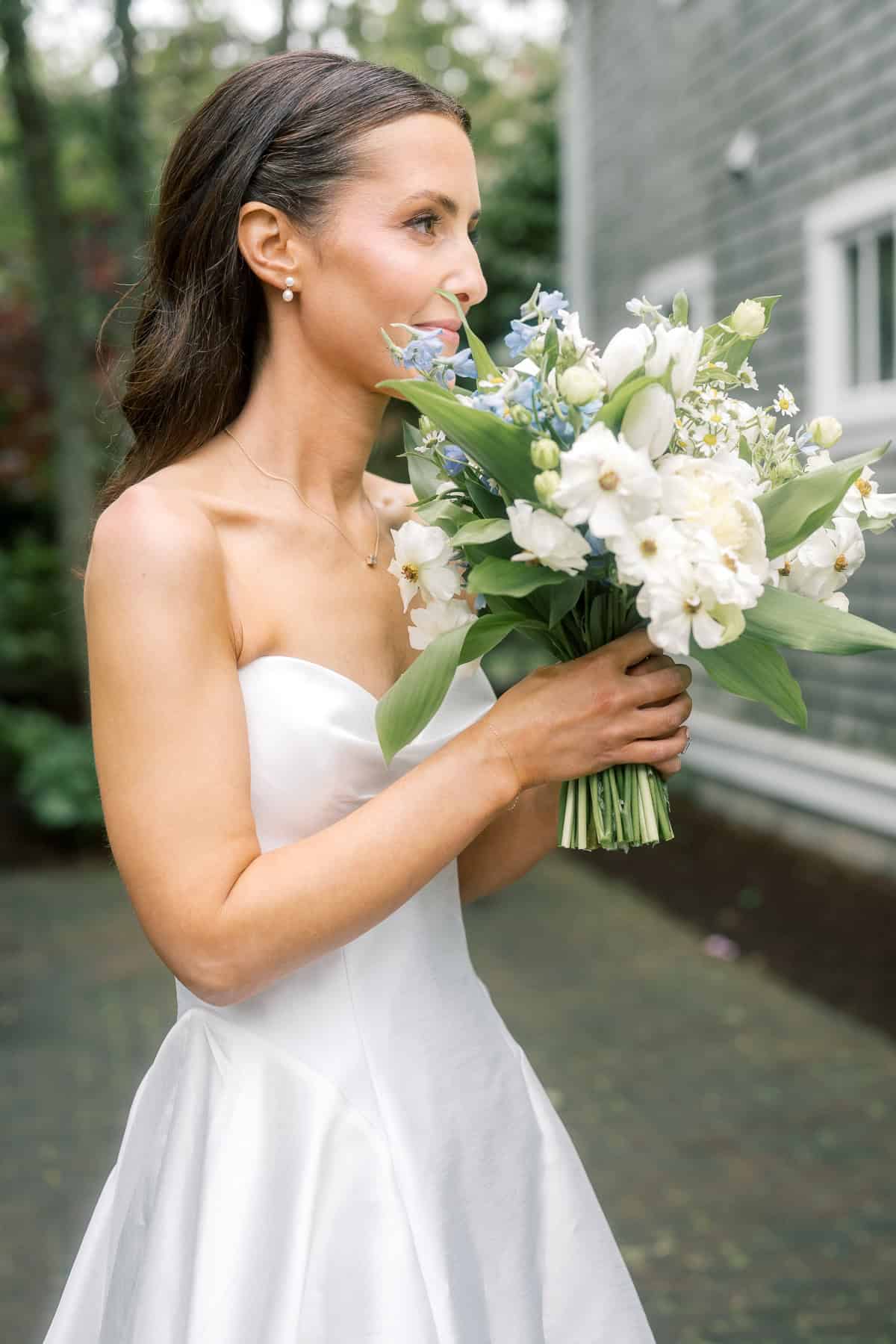 A Nantucket bride in a white strapless dress holds a bouquet of white and light blue flowers outdoors, captured by Nantucket Wedding Photographer Kaity Farrell near a gray building.
