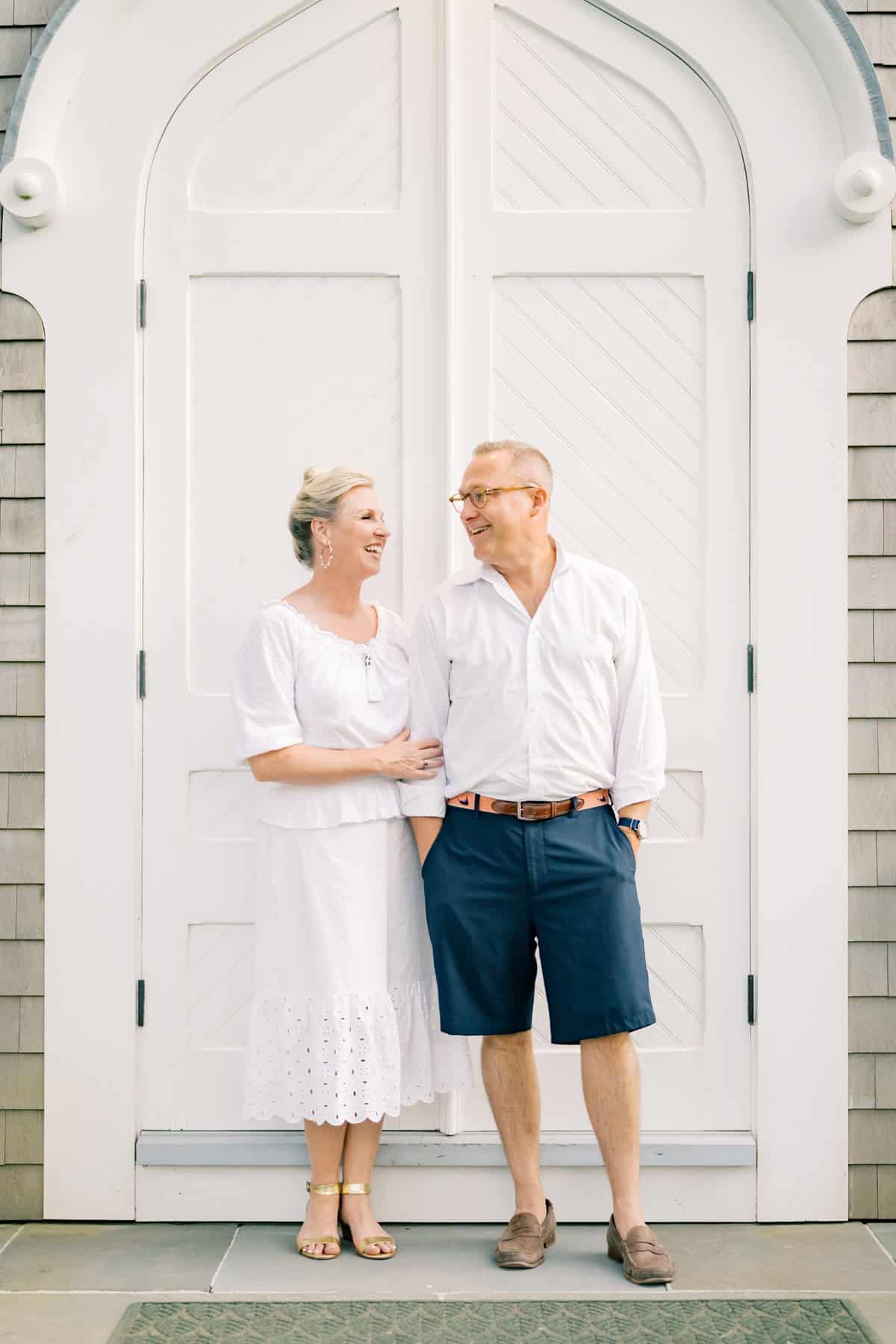 A Nantucket couple stands together smiling in front of large white double doors, both dressed in light, summery clothing—captured beautifully by Nantucket couples photographer Kaity Farrell.
