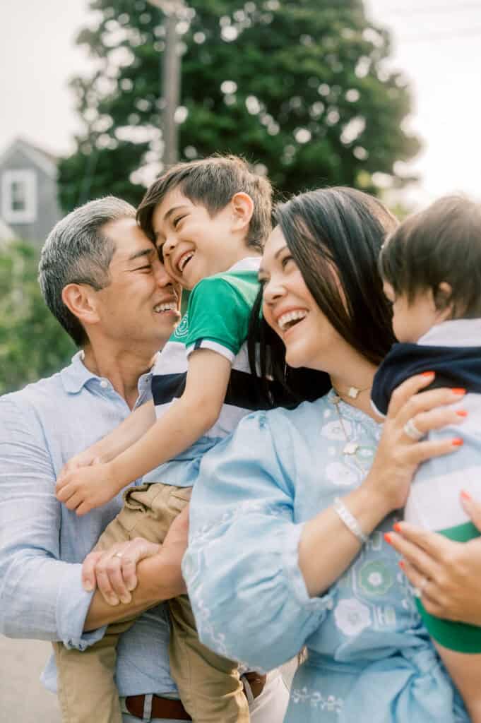 A family of four stands outdoors, smiling and laughing. Captured by Nantucket family photographer Kaity Farrell, the father holds an older son while the mother holds a younger child, with trees and a house in the background.
