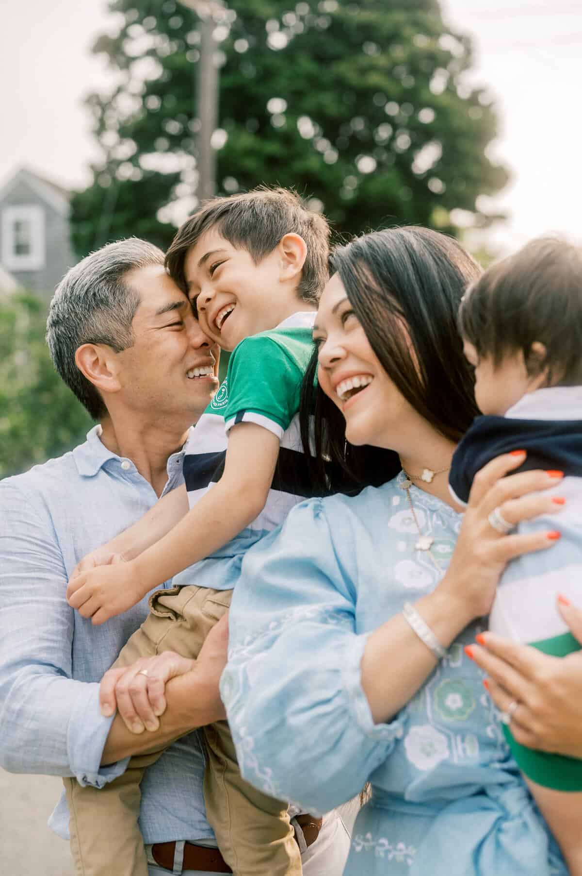 A family of four stands outdoors, smiling and laughing. Captured by Nantucket family photographer Kaity Farrell, the father holds an older son while the mother holds a younger child, with trees and a house in the background.