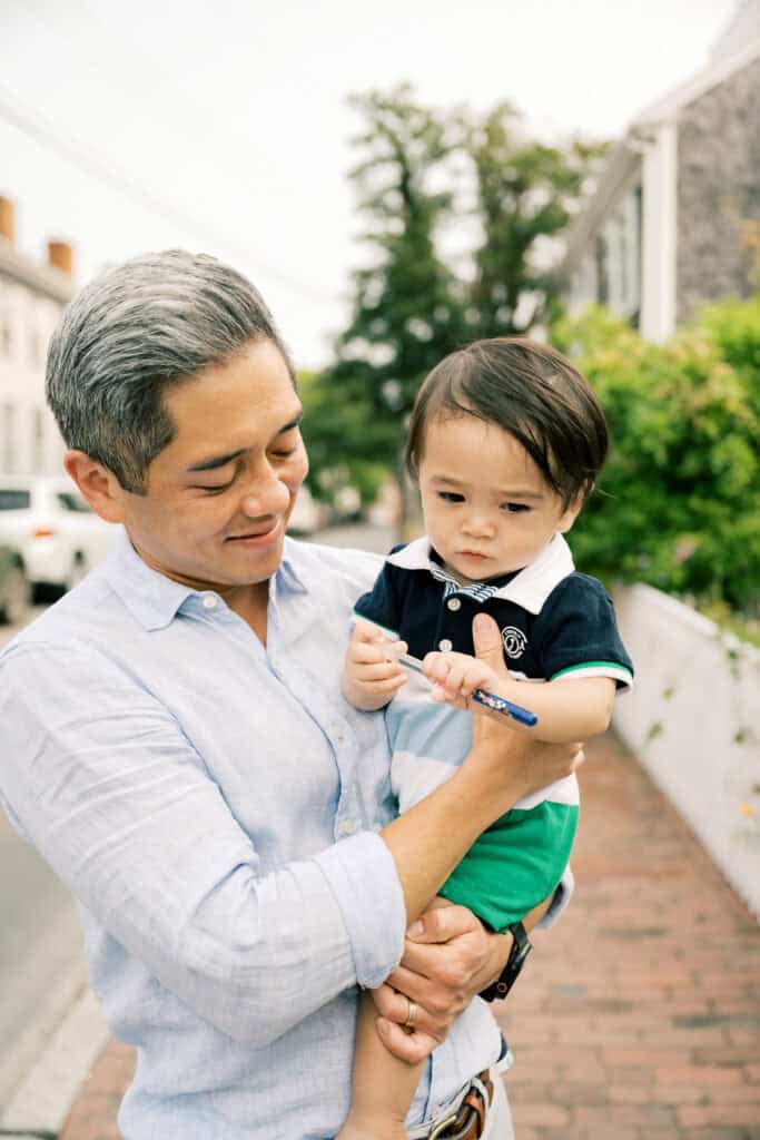 An father holding his young son outdoors on a sidewalk; the child is examining a pen while the adult looks on and smiles during a family portrait session with Nantucket photographer Kaity Farrell.