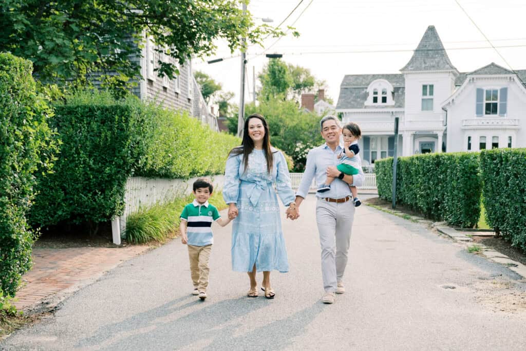 A Nantucket family walks hand-in-hand down a residential street, smiling amid houses and greenery—perfect for a candid family photo session with Nantucket photographer Kaity Farrell.