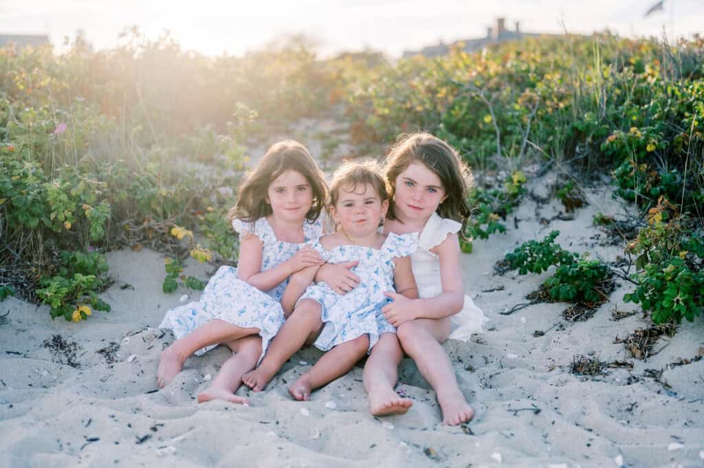 Three young children in light-colored summer dresses sit barefoot on sandy ground surrounded by greenery, with sunlight in the background—capturing the timeless feel of Nantucket beach family photos by photographer Kaity Farrell.