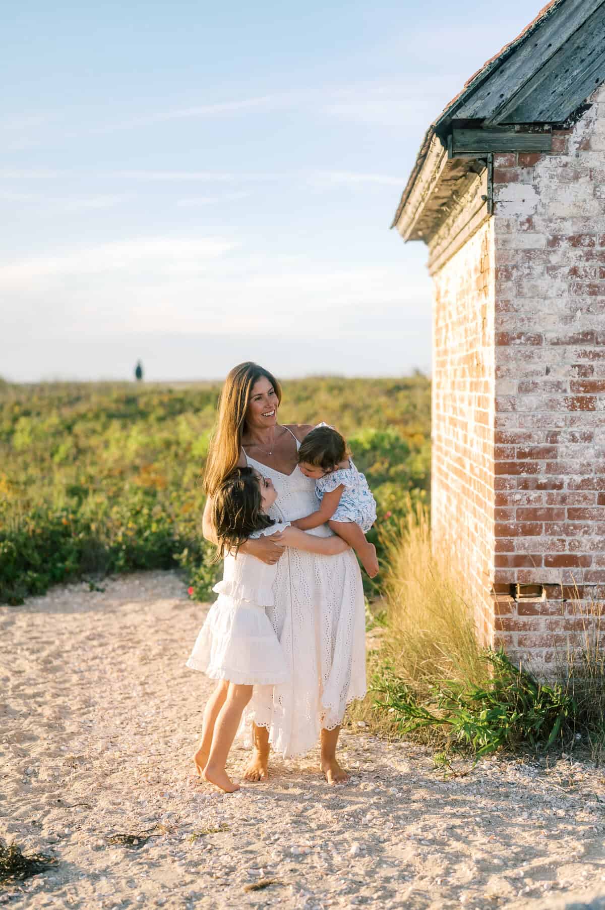 A woman in a white dress stands by a brick building outdoors, hugging two children dressed in light clothing. Grass and sky are visible in the background, capturing a sweet Nantucket family moment during their photo session.