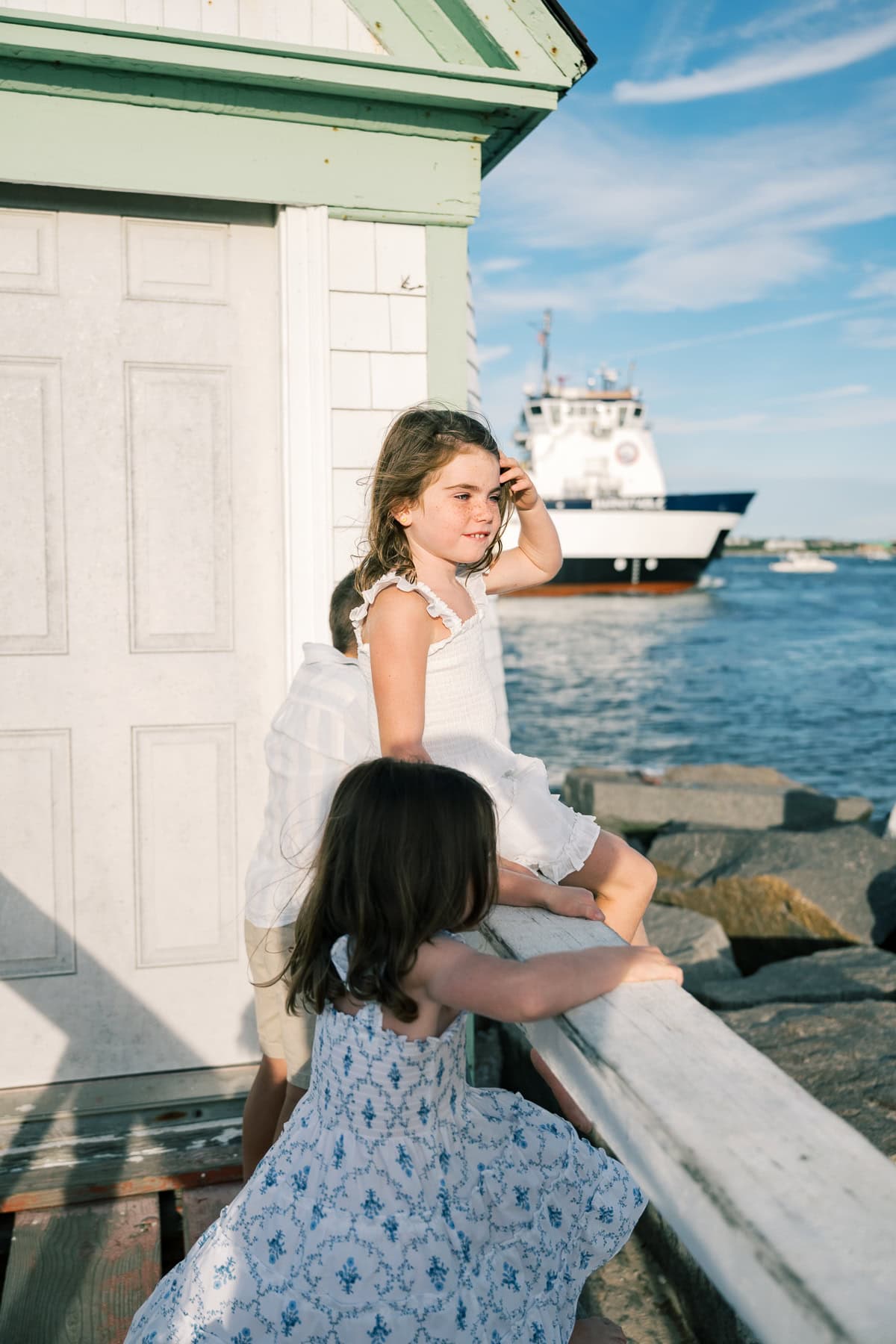 Two young girls in summer dresses stand by a white building near the water, captured by photographer Kaity Farrell, as a boat passes by in the background under a blue sky.