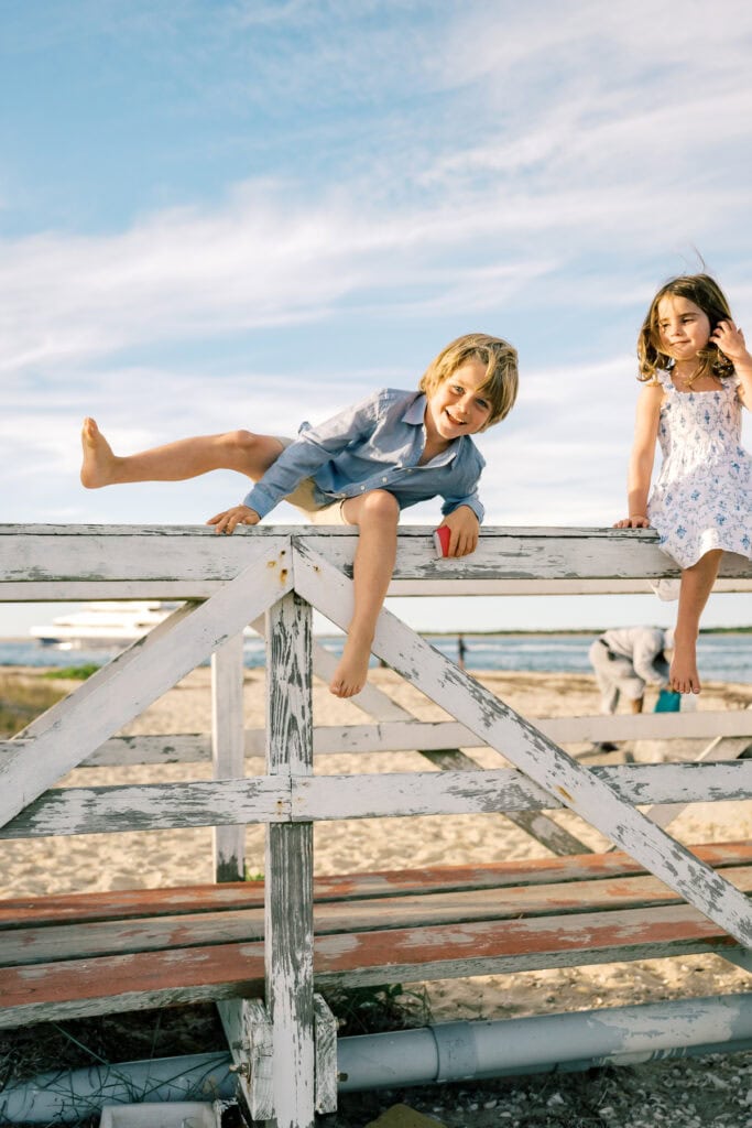 Two children sit and climb on a weathered wooden fence at a beach, with sand and water in the background under a blue sky—captured by photographer Kaity Farrell for Nantucket summer family photos.