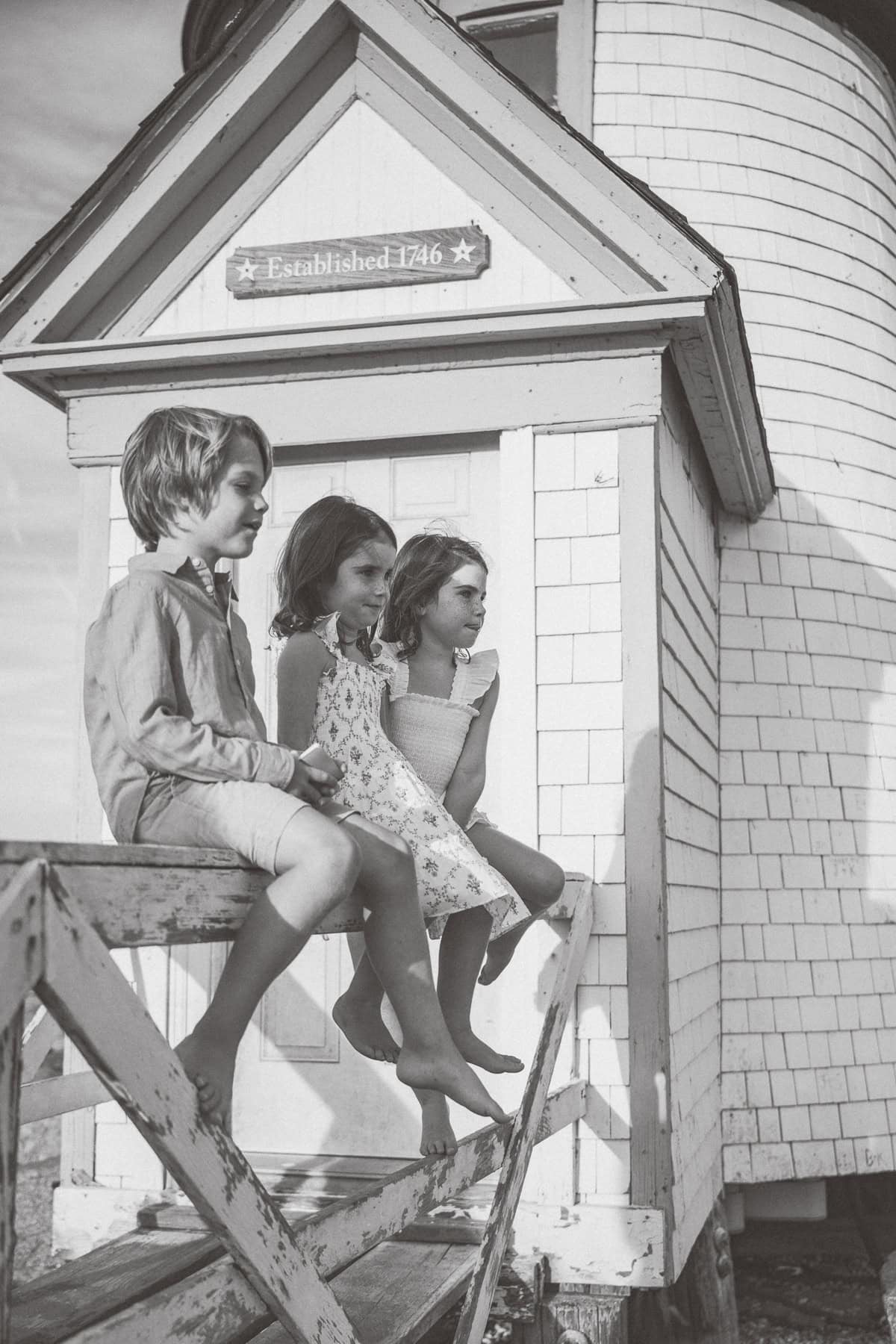 Three children sit on a wooden railing in front of a building labeled "Established 1746," captured beautifully by Nantucket family photographer Kaity Farrell.