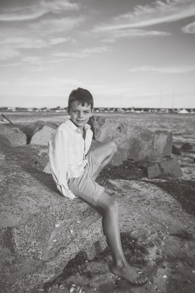 A boy in a light shirt and shorts sits barefoot on large rocks by the water, with sailboats and a cloudy sky behind him. Captured in black and white by photographer Kaity Farrell, it evokes timeless Nantucket vacation family photos.