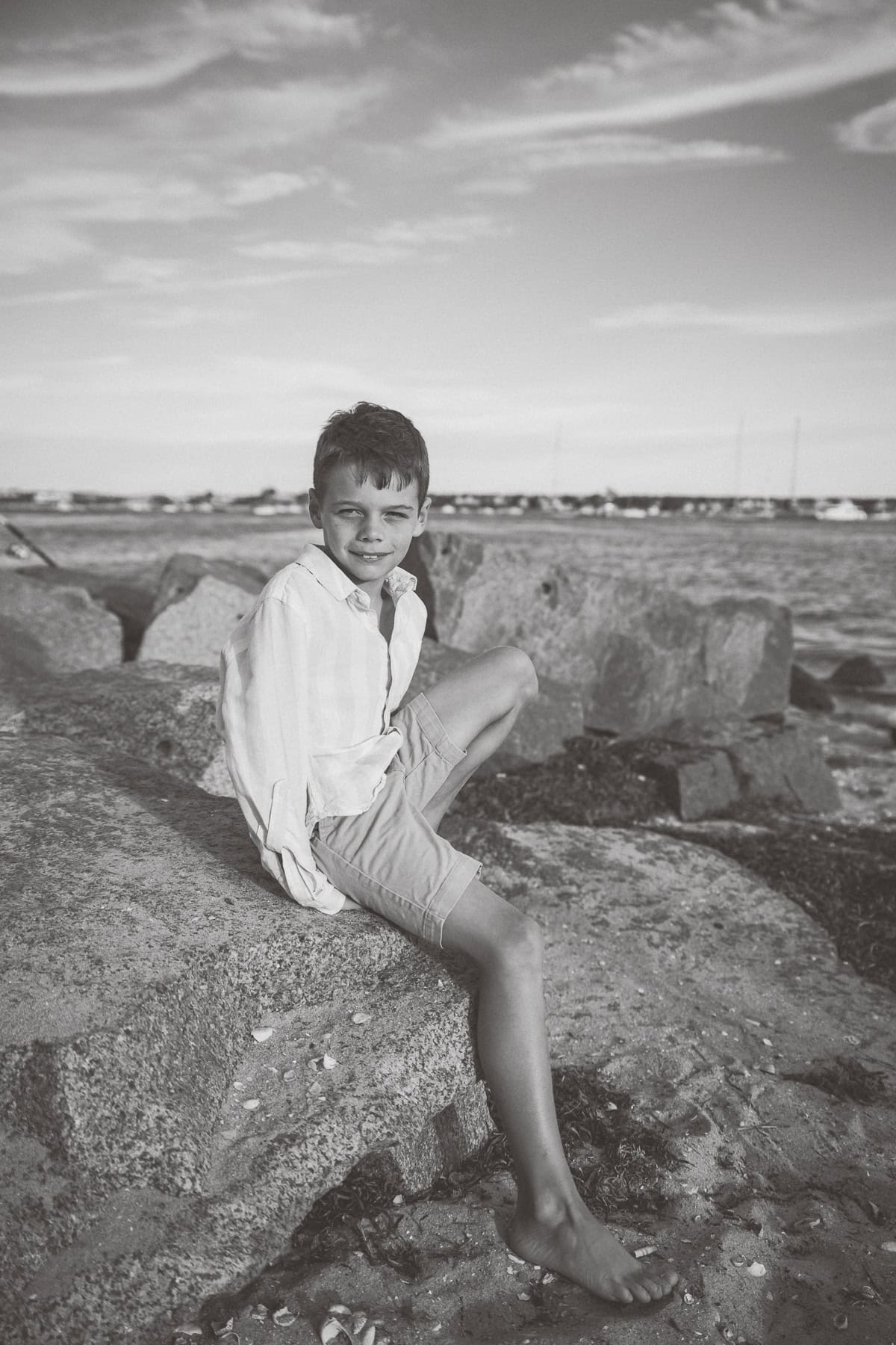 A boy in a light shirt and shorts sits barefoot on large rocks by the water, with sailboats and a cloudy sky behind him. Captured in black and white by photographer Kaity Farrell, it evokes timeless Nantucket vacation family photos.