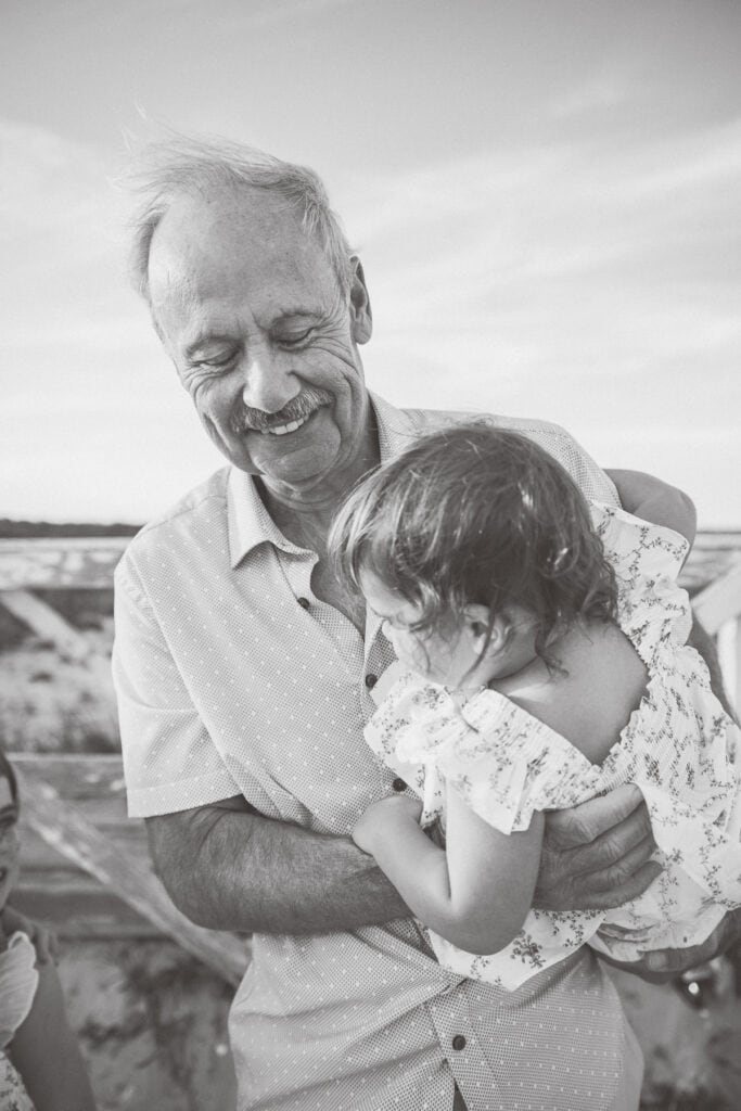 An older man smiles while holding a young child outdoors during a Nantucket extended family portrait session. Both wear light-colored clothing in this black and white photo by photographer Kaity Farrell.