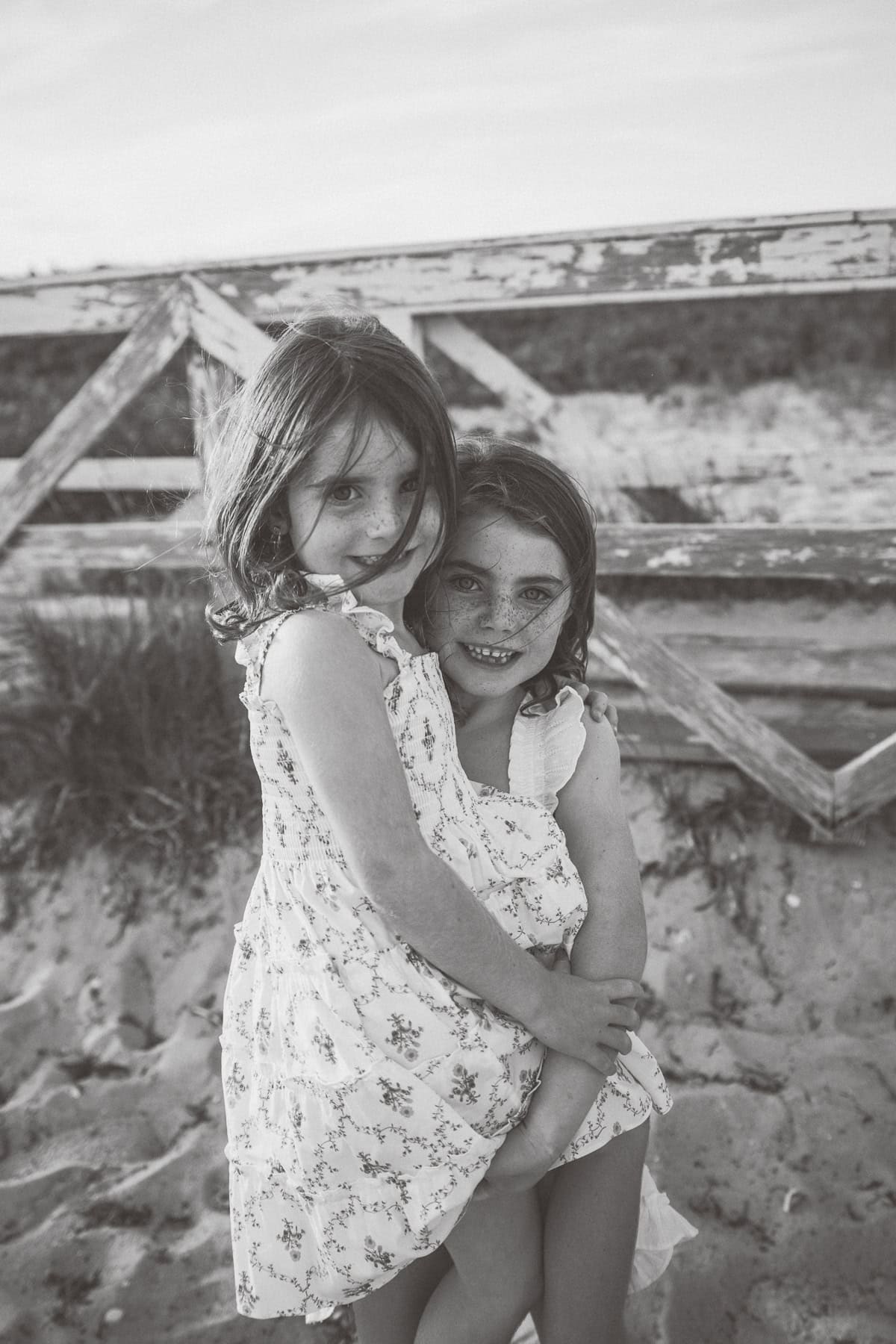 Two young girls in floral dresses stand close together on a sandy beach, with one sitting and the other standing in front of a weathered wooden fence during a Nantucket family photo session.