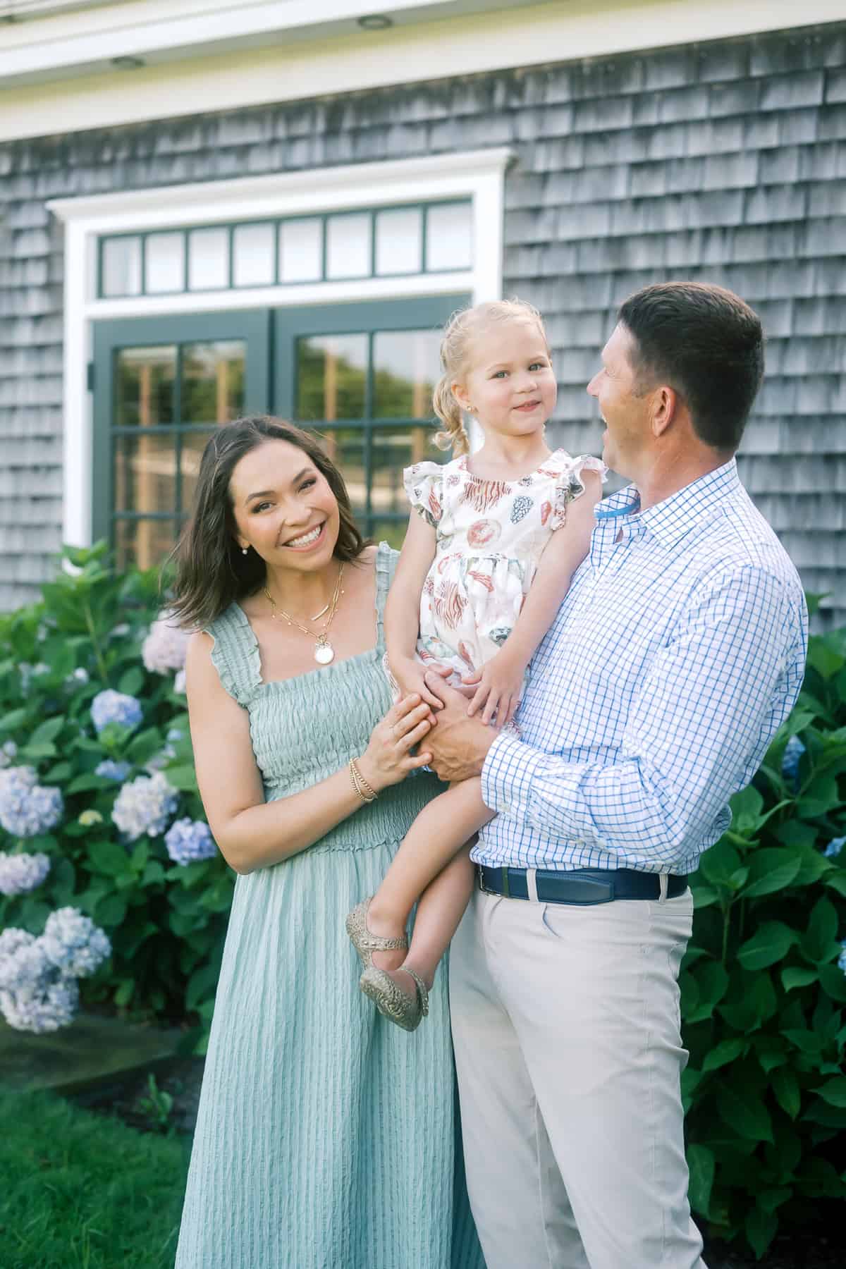A woman, man, and young girl pose smiling in front of a house with hydrangea bushes, the man holding the girl in his arms—captured beautifully by Nantucket family photographer Kaity Farrell.
