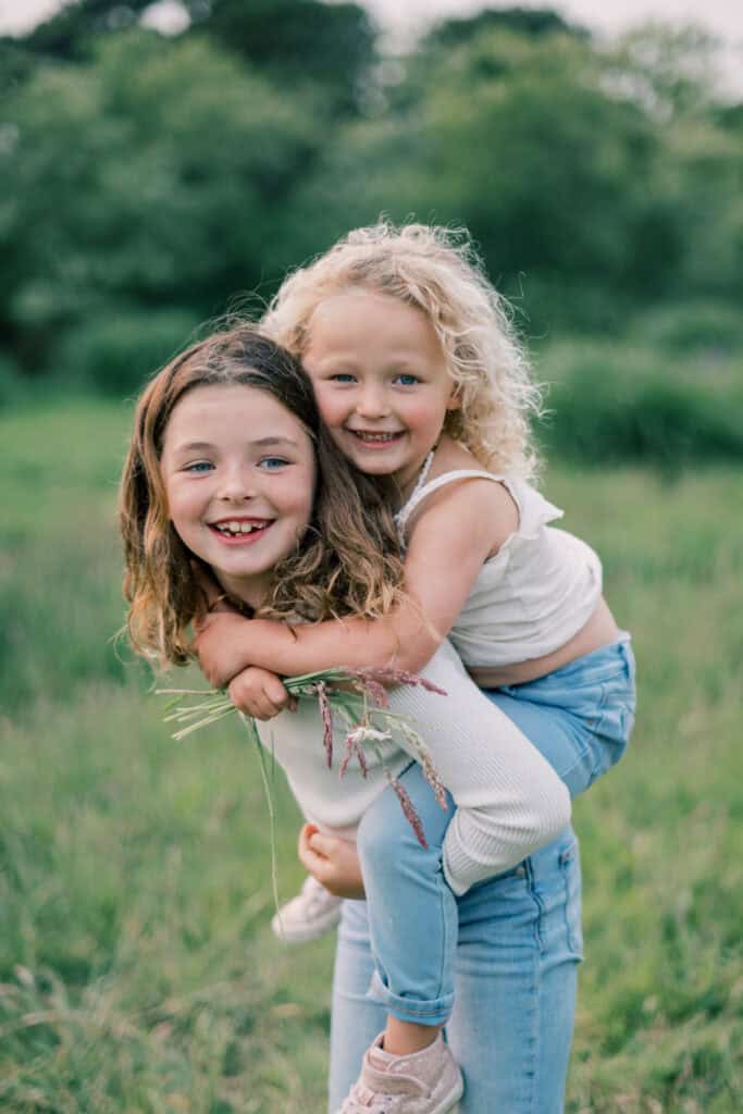 Two young girls enjoy a playful moment in a grassy field during a Nantucket family photo session. One gives the other a piggyback ride as they both smile at the camera, perfectly capturing memories with Nantucket photographer Kaity Farrell.