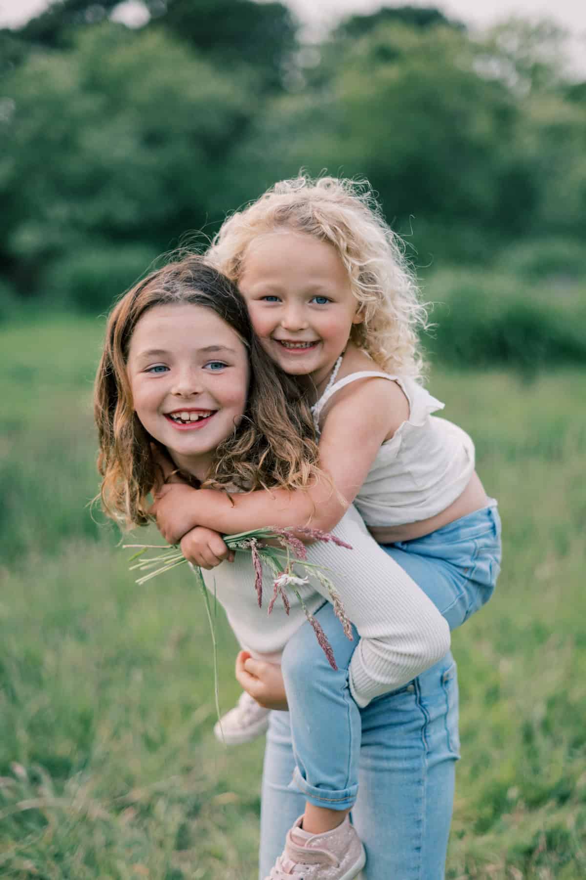 Two young girls enjoy a playful moment in a grassy field during a Nantucket family photo session. One gives the other a piggyback ride as they both smile at the camera, perfectly capturing memories with Nantucket photographer Kaity Farrell.