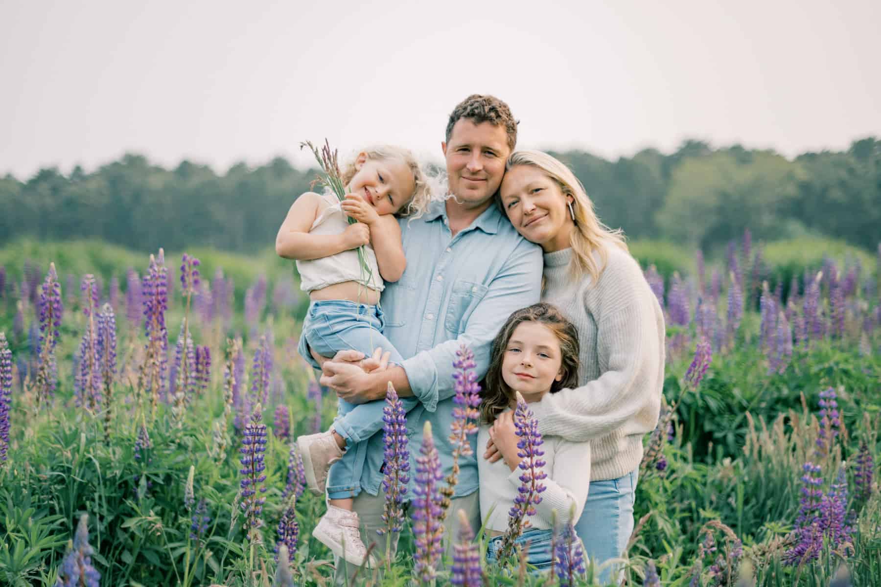 A family of four poses together in a field of purple lupine flowers, with two children and two adults standing close and smiling at the camera, beautifully captured by Nantucket portrait photographer Kaity Farrell.