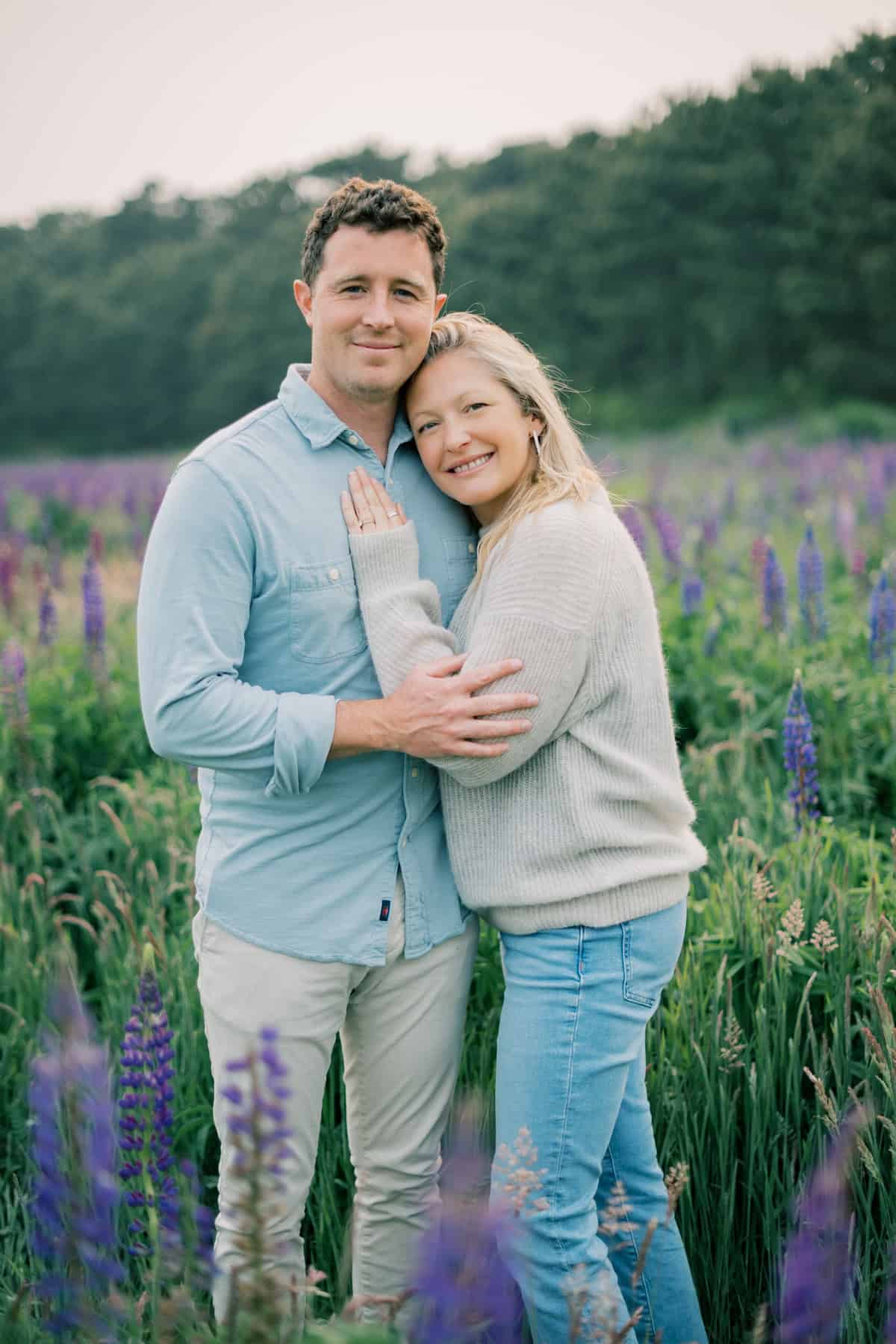 A couple stands close together in a field of purple flowers, smiling at the camera with greenery and trees in the background during a couples photo session with Nantucket photographer Kaity Farrell.
