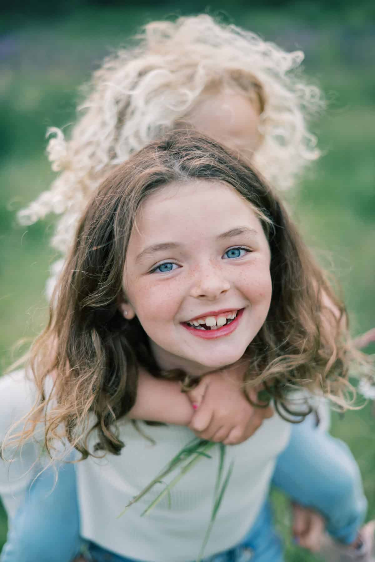 Two children are outdoors on Nantucket; one with curly blonde hair gives a piggyback ride to another child with brown hair and blue eyes, who is smiling at the camera—captured beautifully by Nantucket family photographer Kaity Farrell.