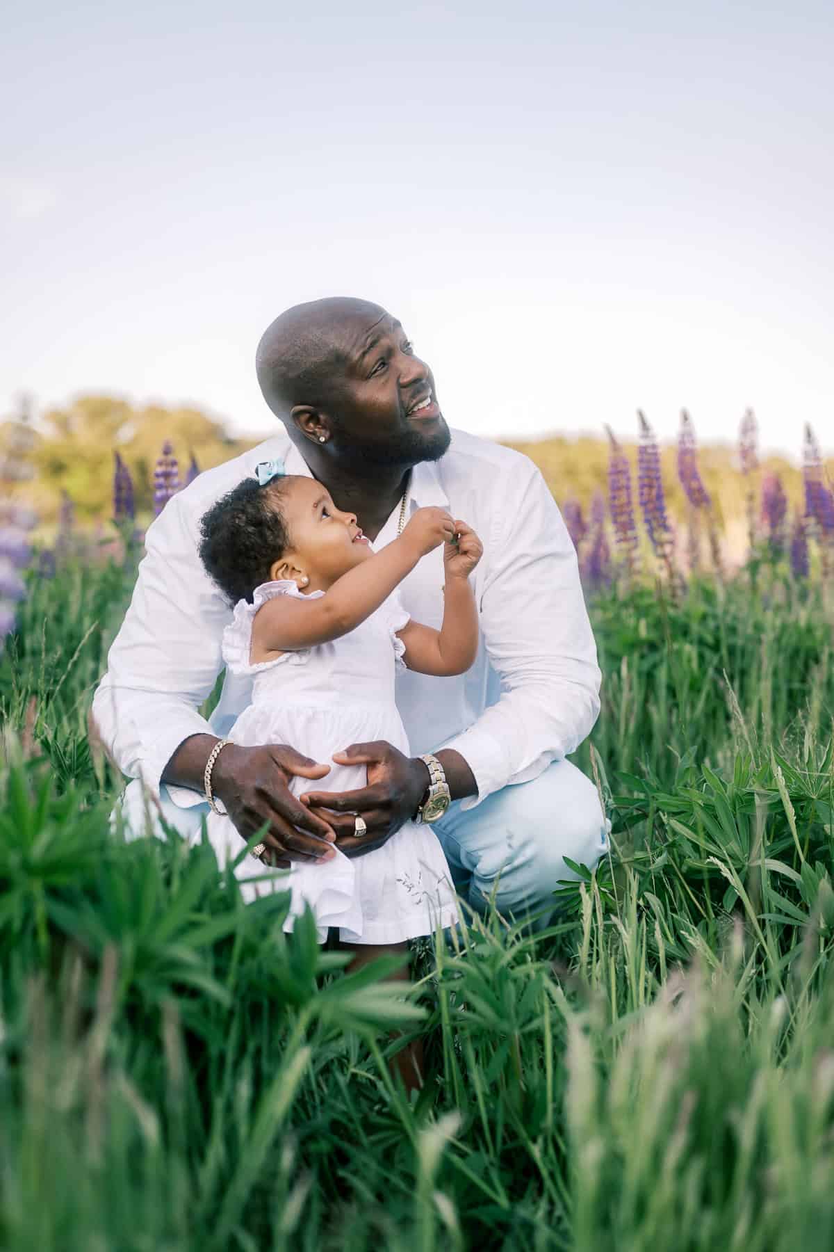 A father and young daughter in white clothes sit together in a field of tall green grass and purple lupine flowers, looking upward—a serene moment perfect for Nantucket family portraits by photographer Kaity Farrell.