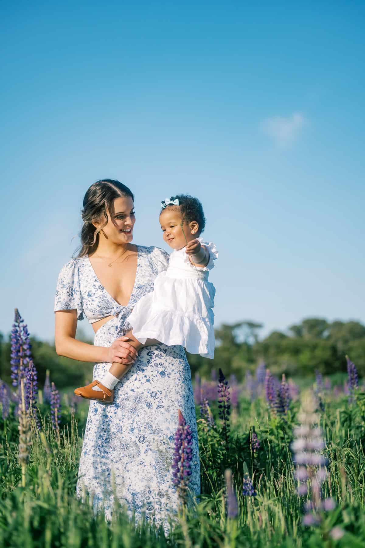 A mother in a floral dress holds her baby girl in a white outfit while standing in a field of purple lupine flowers under a clear blue sky, captured beautifully by Nantucket family photographer Kaity Farrell.