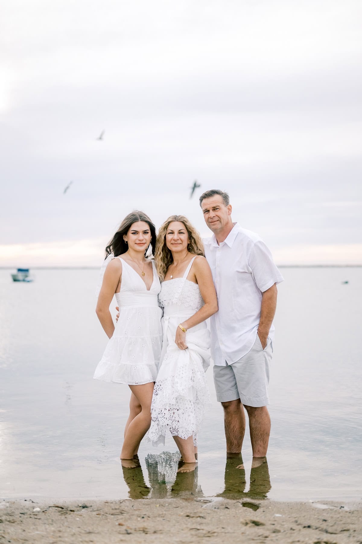 Three adults stand together in shallow water at the beach, dressed in white, with a calm sea, distant boat, and seagulls in the background—a timeless moment captured by Nantucket portrait photographer Kaity Farrell.