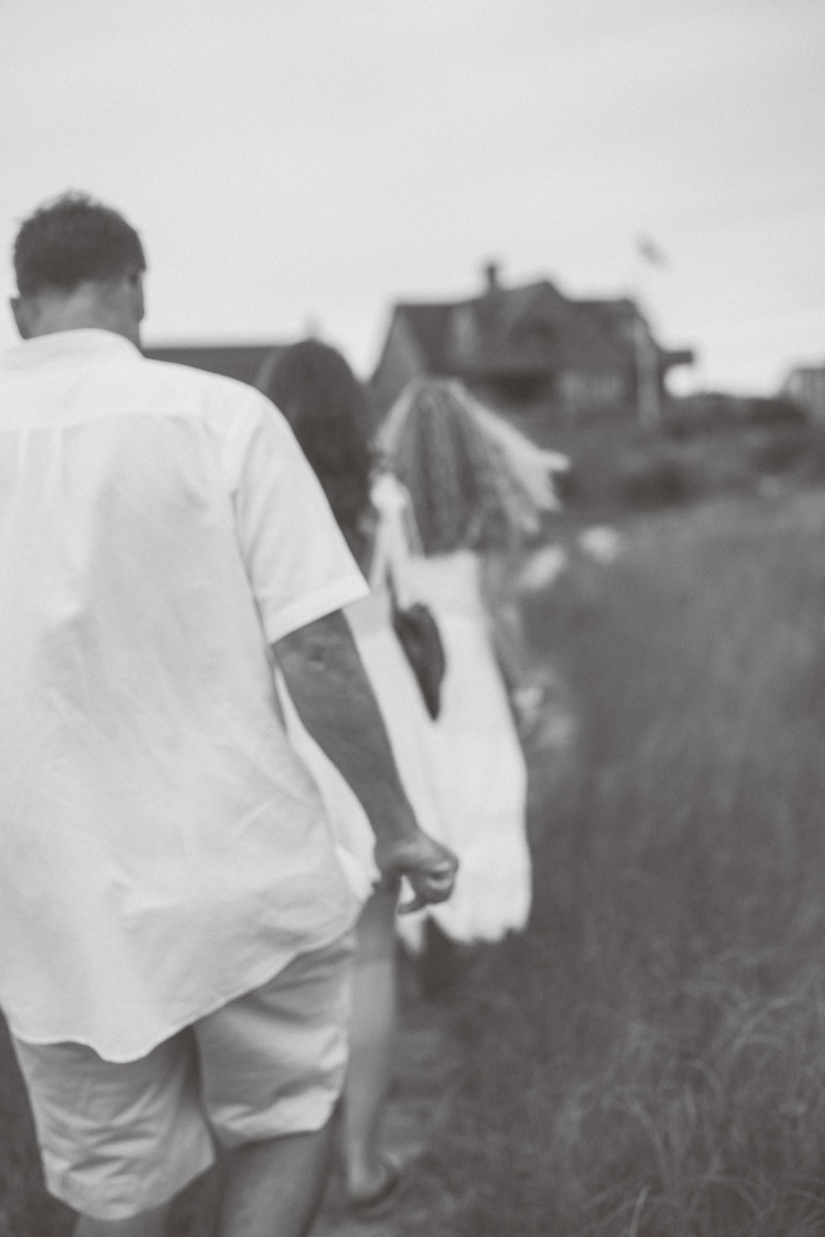 A group of people in light-colored clothing walk in a line along a grassy path toward houses, photographed from behind in black and white during a family photo session with Nantucket photographer Kaity Farrell.