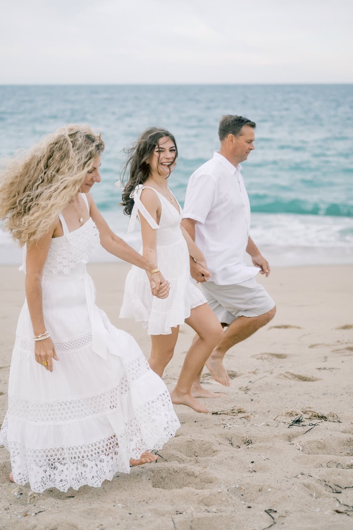 Three people dressed in white walk hand-in-hand along a sandy beach near the water on an overcast day during a Nantucket family photo session with Nantucket photographer Kaity Farrell.