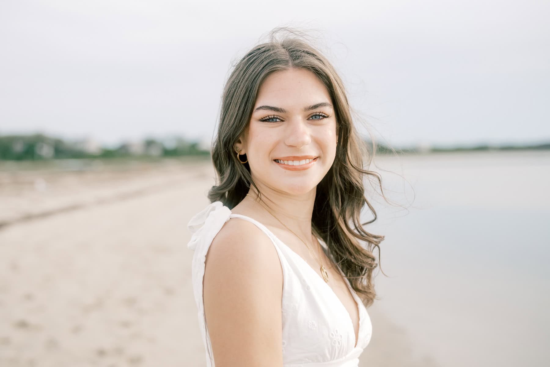 A young woman with long brown hair in a white dress stands on a sandy beach near calm water, smiling at the camera during her high school senior photo session with Nantucket photographer Kaity Farrell.