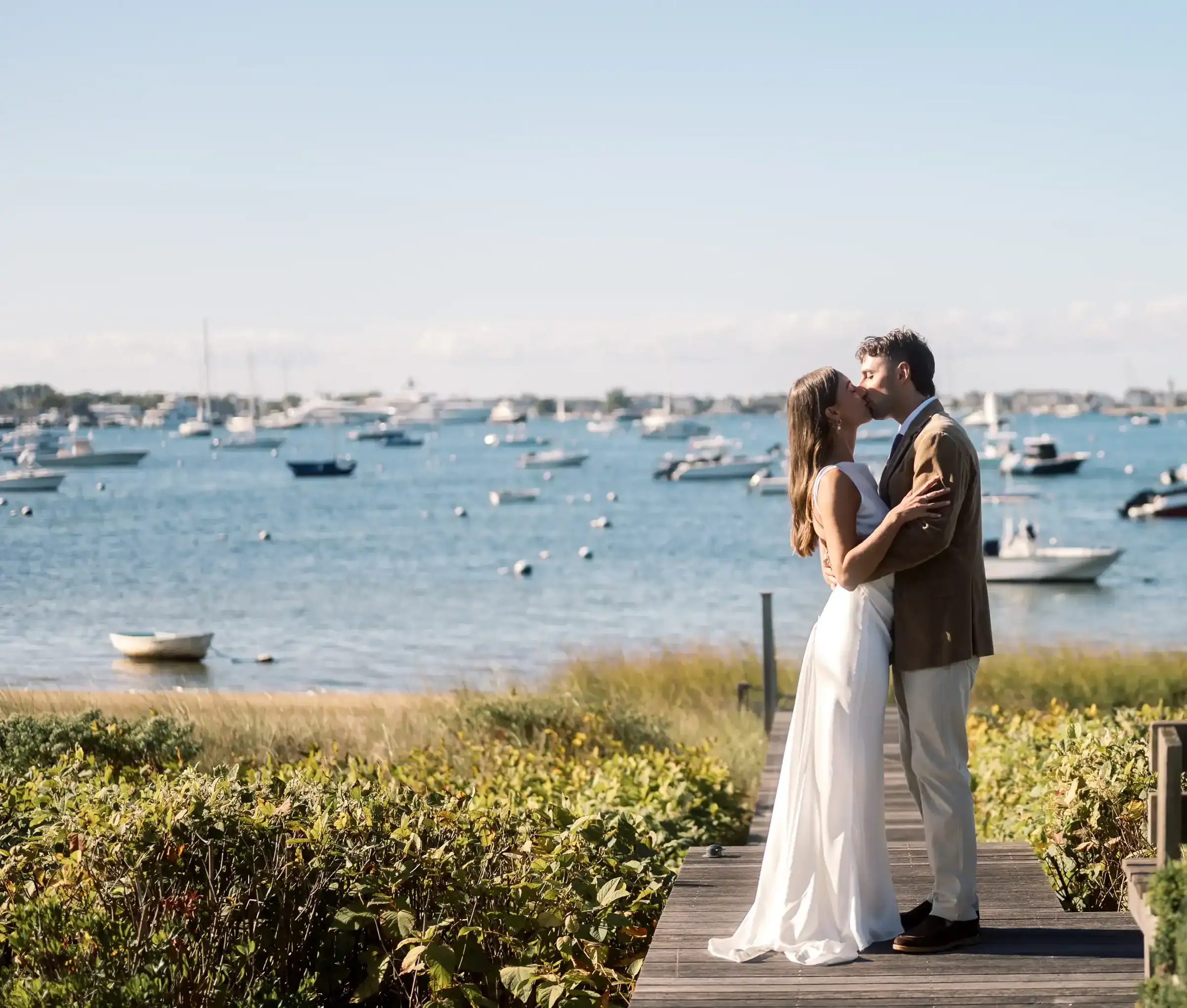 A couple dressed in wedding attire stands on a dock, kissing, with a marina full of boats and a clear blue sky in the background.