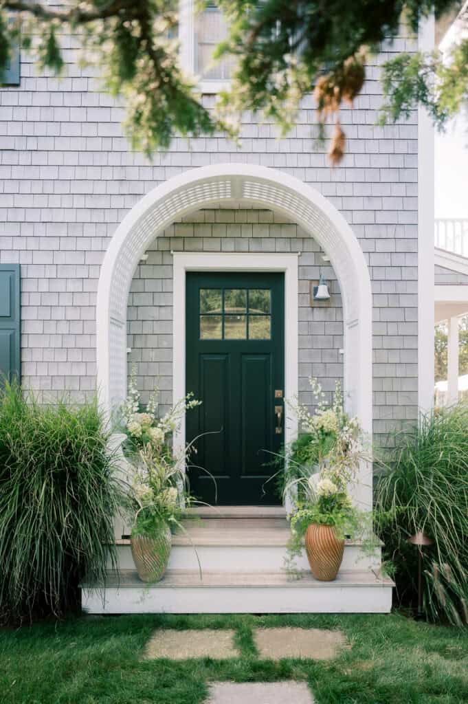 A dark green front door with a window is framed by a white arched trellis at this private residence in Monomoy. Two large planters with green foliage flank the entrance, and ornamental grasses line the pathway leading to the steps.
