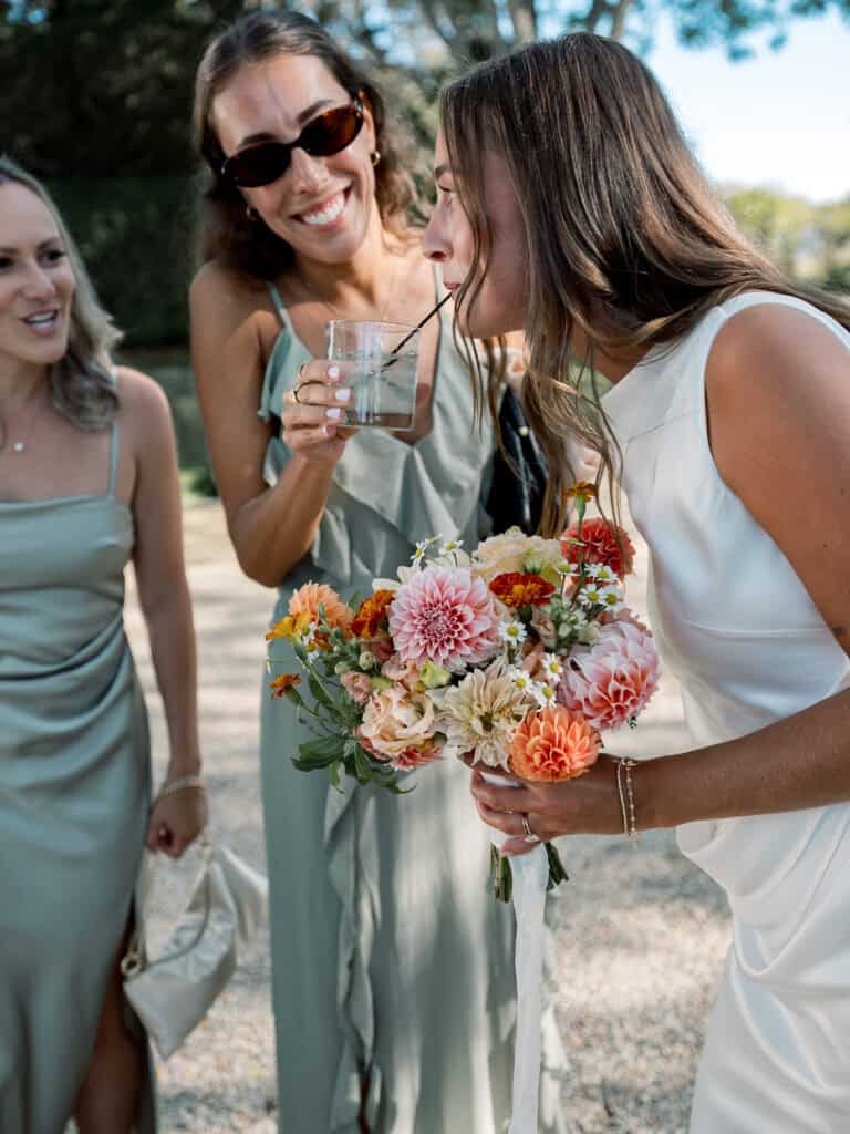 A nantucket bride in a white dress holding a colorful bouquet sips a drink through a straw held by a smiling friend in sunglasses; another woman in a light green dress stands nearby outdoors.