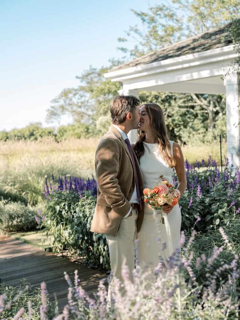 A couple stands close together in a garden near a white gazebo, the woman holding a bouquet of flowers. They are dressed formally and smiling, perfectly captured by a Nantucket wedding photographer.
