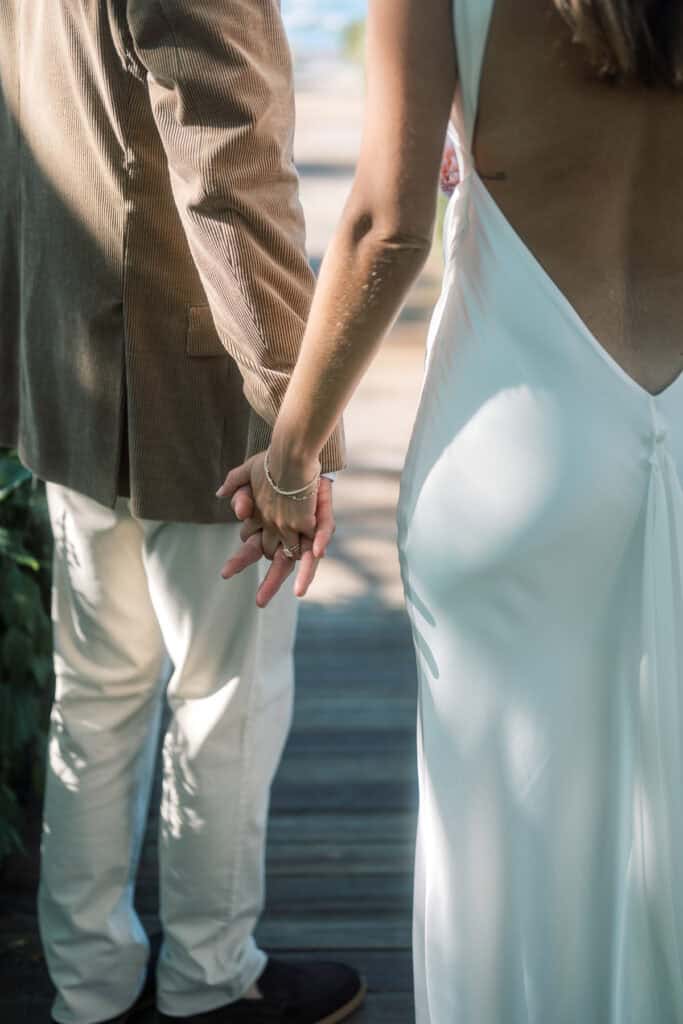 A bride and groom hold hands side by side. The woman wears a white, backless dress and the man a brown jacket with light pants. Sunlight softly illuminates their hands and clothing, capturing this intimate Nantucket wedding moment.