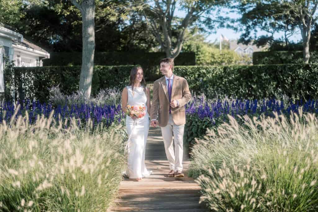 A Nantucket bride in a white dress holding a bouquet and her groom in a tan jacket and tie walk hand in hand on a garden path surrounded by tall grasses and purple flowers on a sunny day.
