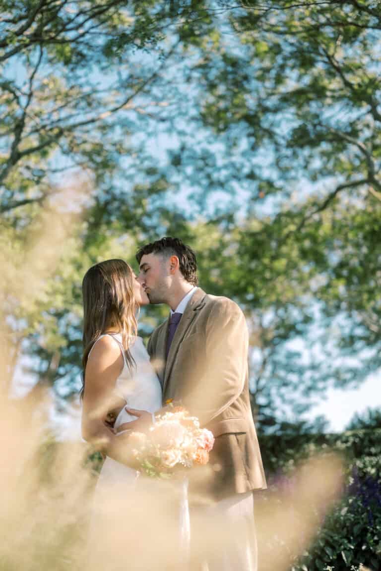 A couple stands outdoors, embracing and about to kiss, surrounded by trees and sunlight. Captured by a Nantucket wedding photographer, the woman holds a bouquet of flowers; both are dressed formally, with soft focus plants in the foreground.