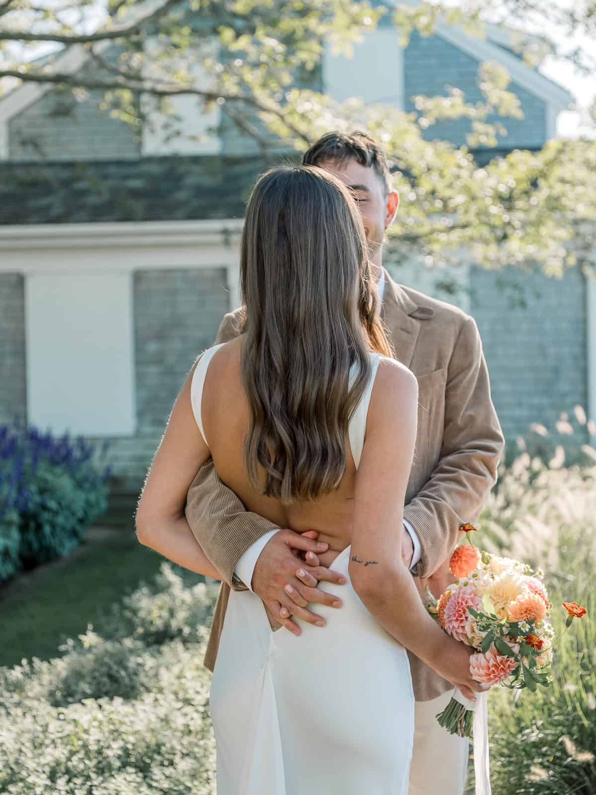 A bride and groom share an intimate embrace outdoors for a Nantucket wedding portrait, with the bride in a white backless dress and bouquet and the groom in a tan suit, surrounded by lush greenery and a charming house.